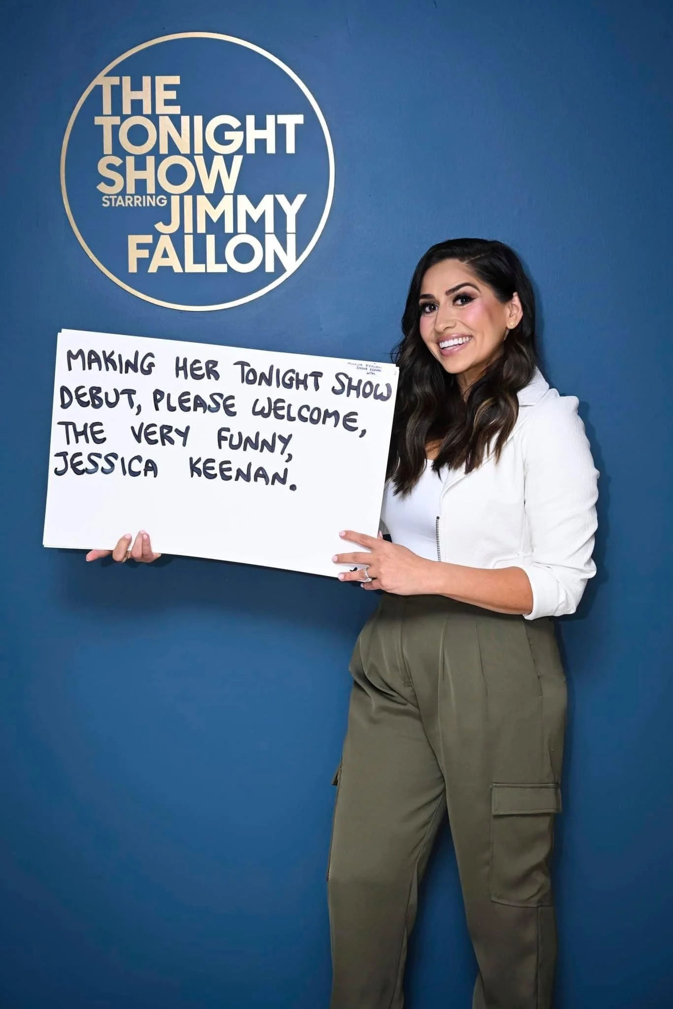 A woman with dark hair, smiling and wearing a white jacket and khaki pants, holding a white sign with black handwritten text in front of a blue wall with the logo of "The Tonight Show Starring Jimmy Fallon".