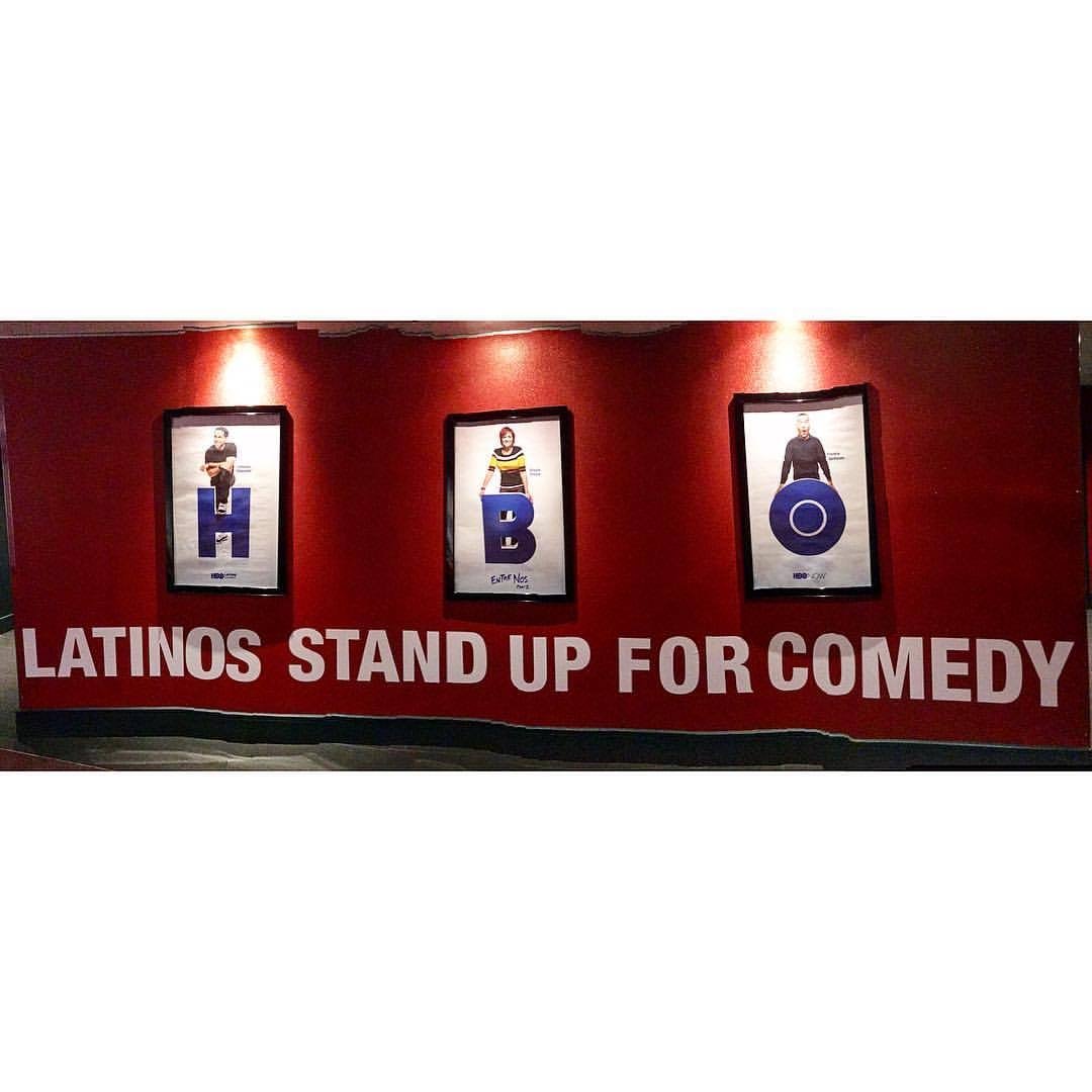 Red wall with three framed posters of people holding large letters S, B, O, and a message that reads "Latinos Stand Up for Comedy."