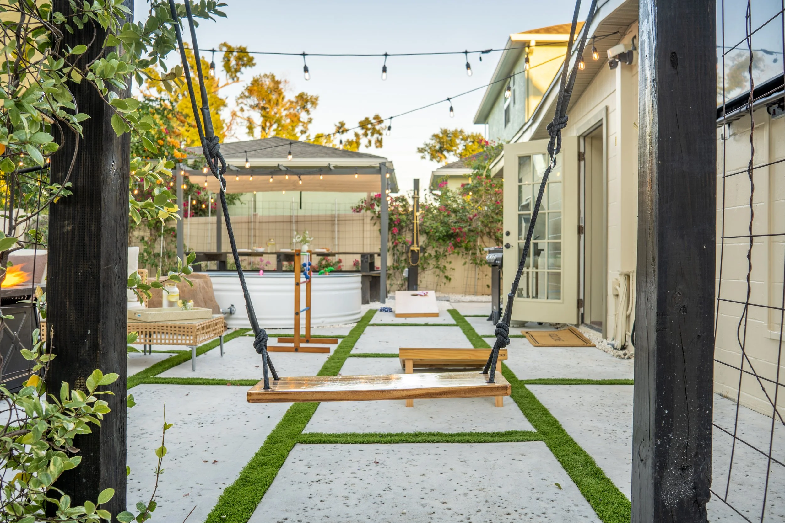 A backyard with string lights, a play area, a small above-ground pool, a swing, and various outdoor furniture and plants, surrounded by trees and a fence.