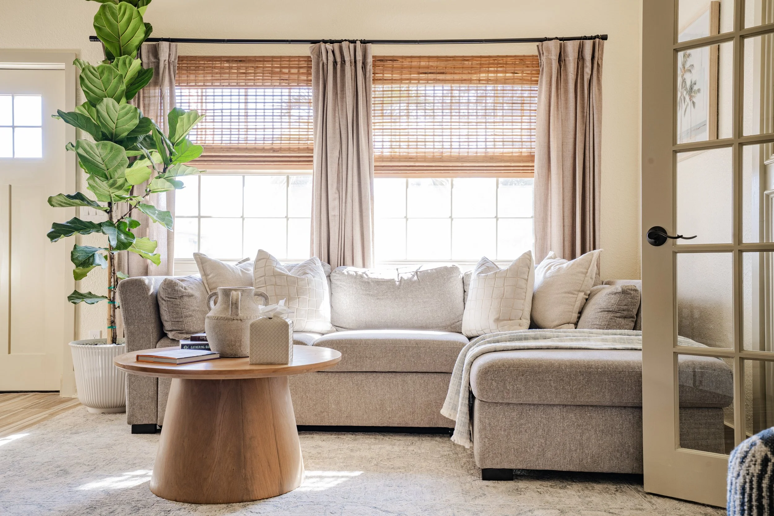 A cozy living room with a large window covered by wooden blinds and beige curtains, a sectional sofa with white pillows, a round wooden coffee table, a potted fiddle leaf fig plant, and sunlight streaming through the window.
