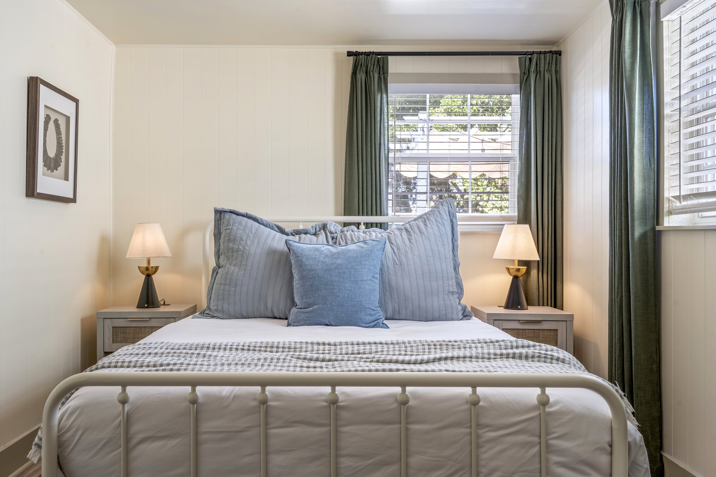 A bedroom with a white metal bed frame, two gray pillows, and matching bedside tables with black lamps. There are large windows with green curtains and blinds, letting in natural light, and a piece of abstract artwork on the white paneled walls.