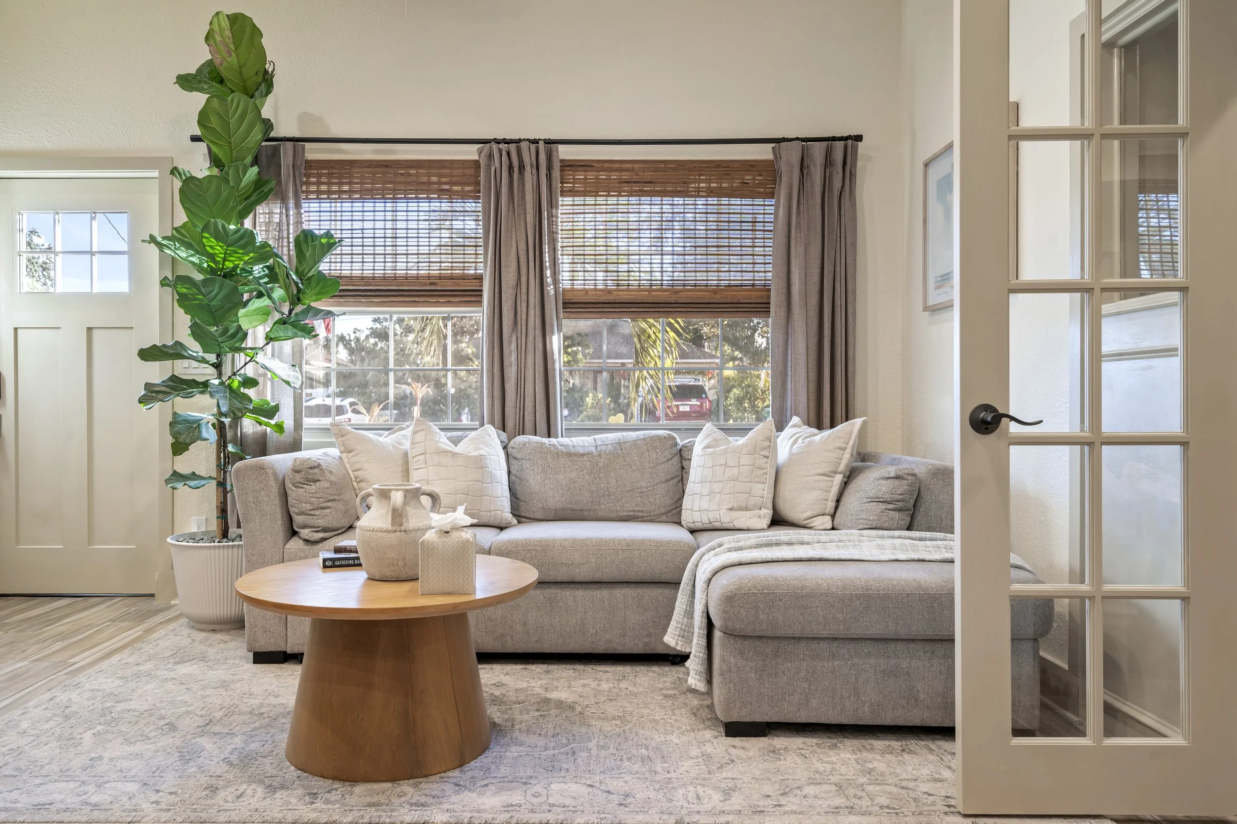 Living room with beige sofa, white pillows, wooden coffee table, greenery, large window with blinds and curtains, view of outdoor trees and house.