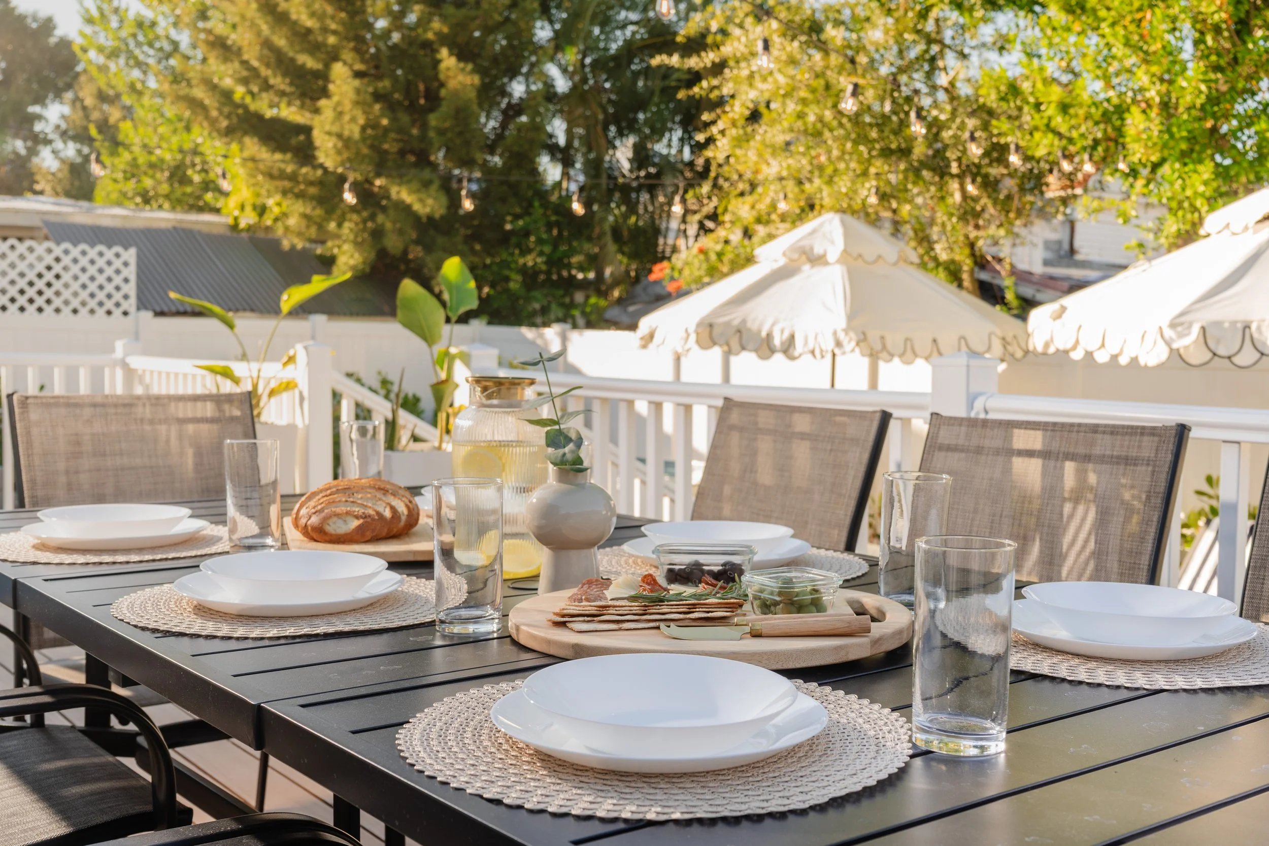 A dining table set outdoors with plates, glasses of water, a loaf of bread, and a small bowl of olives. In the background, there are white patio umbrellas, a white fence, and trees with green and yellow leaves. String lights are also visible hanging 