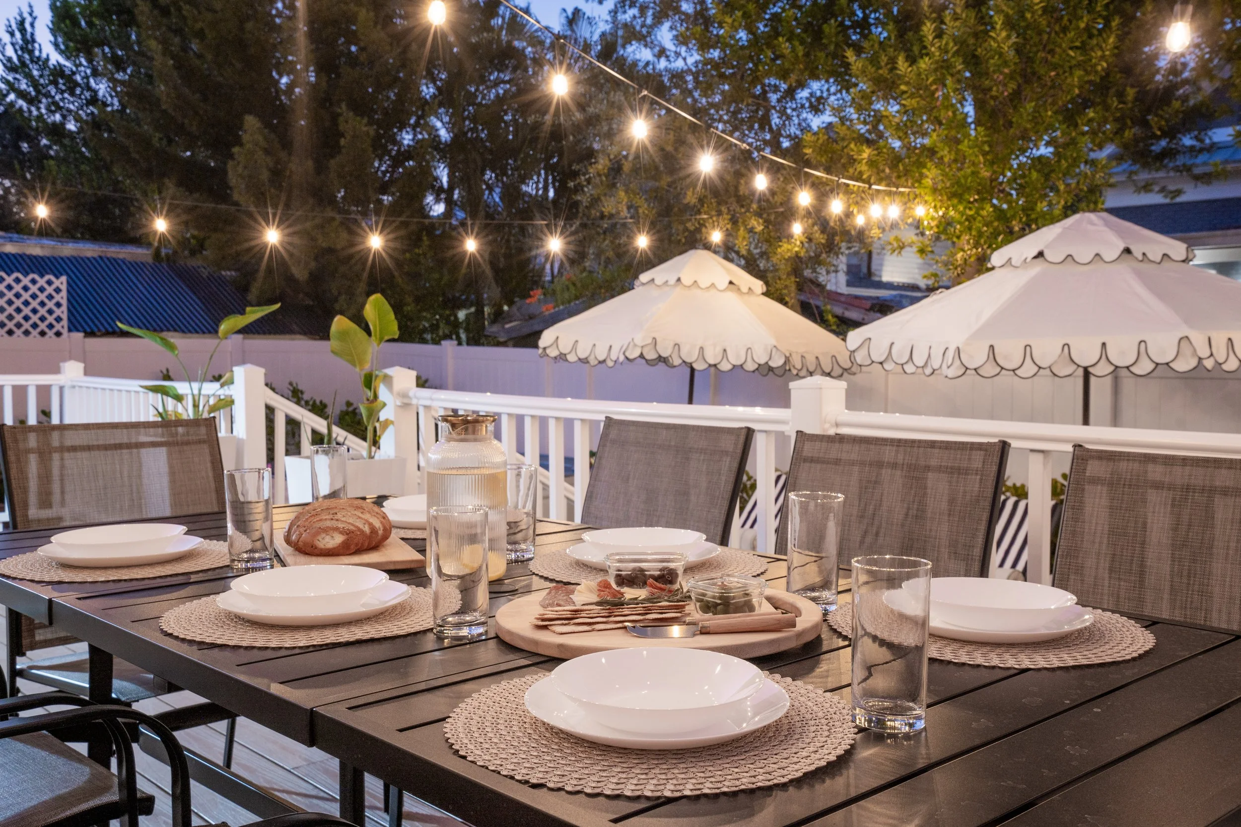 A backyard patio setup at dusk with string lights overhead, large umbrellas, a white fence, and a table set with plates, glasses, bread, and charcuterie.