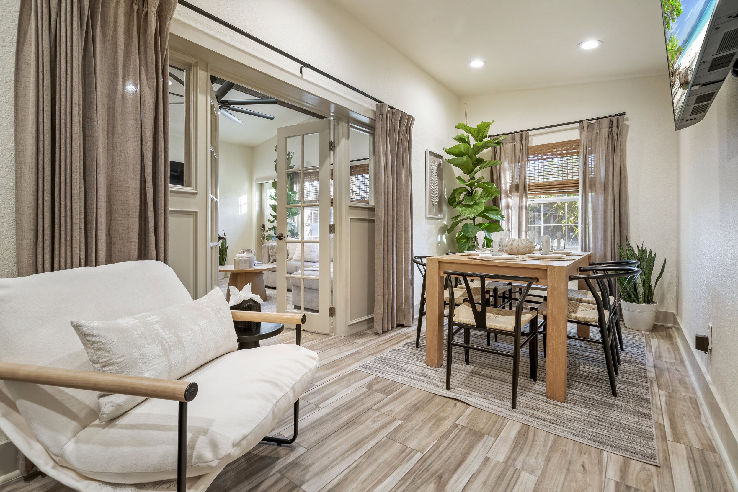 Interior of a living room with white walls, beige curtains, a white sofa, a wooden dining table with black chairs, indoor plants, and large windows allowing natural light.