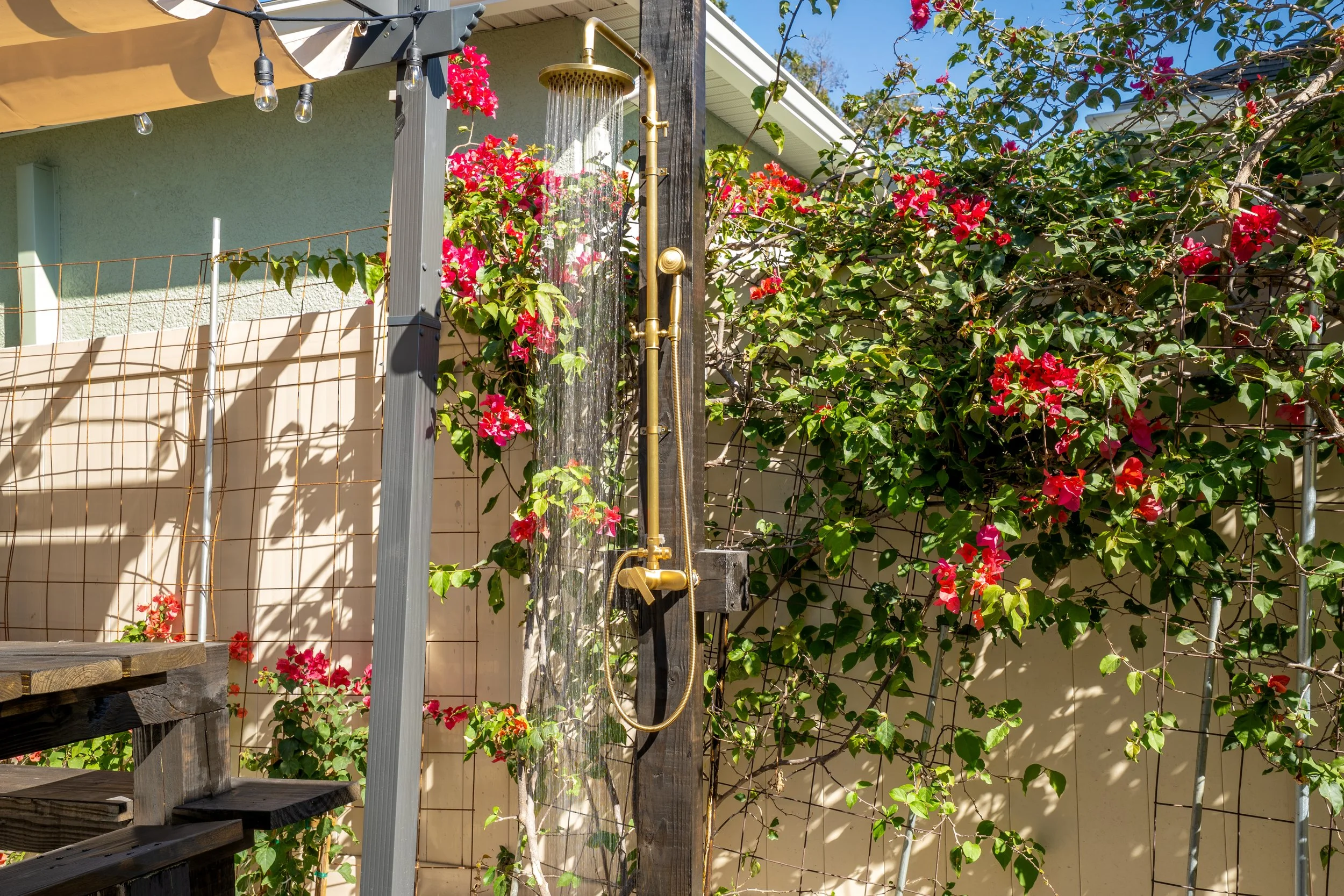 Outdoor shower with brass fixtures mounted on wooden post, surrounded by pink flowering plants and a garden fence, in a sunny backyard.