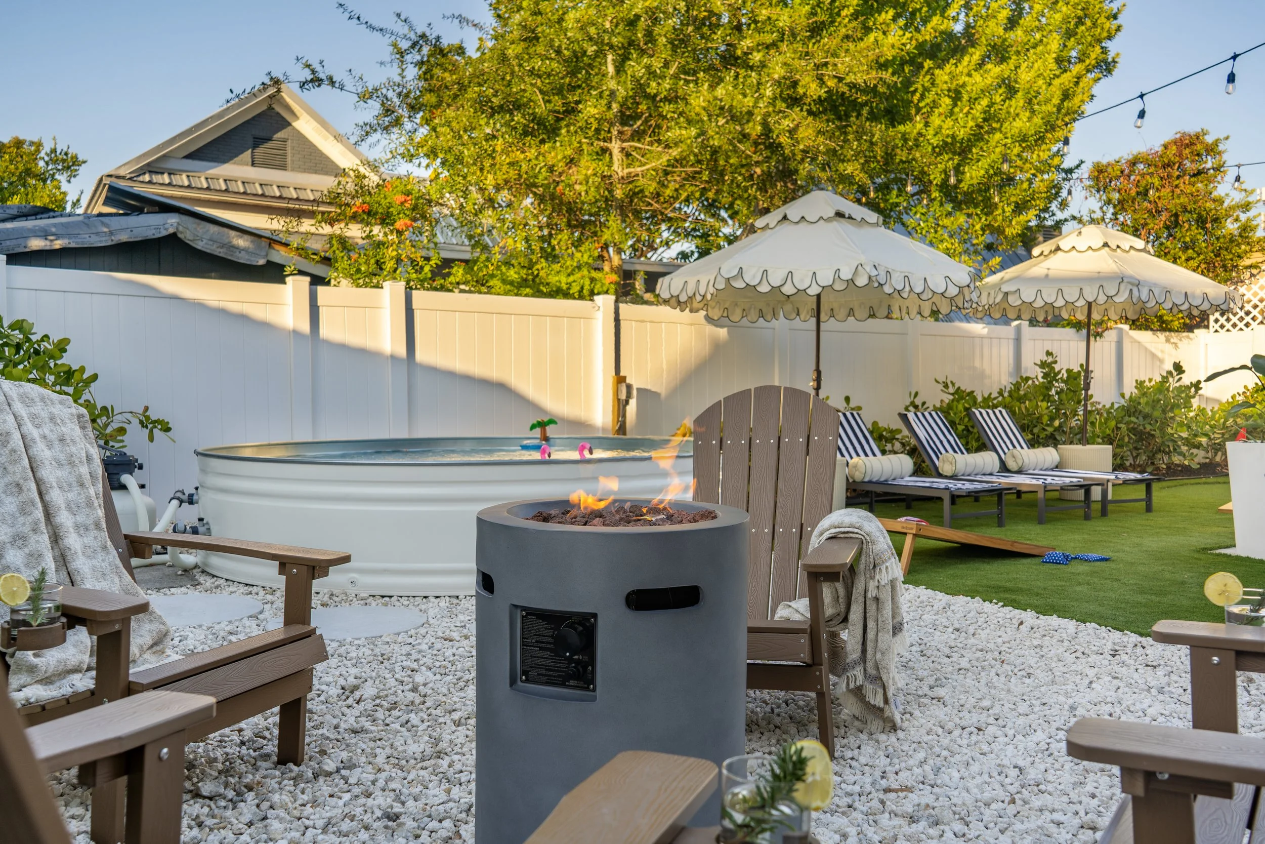 Backyard with lounge chairs, umbrellas, a fire pit, a small pool, string lights, trees, and a white fence under clear blue sky.