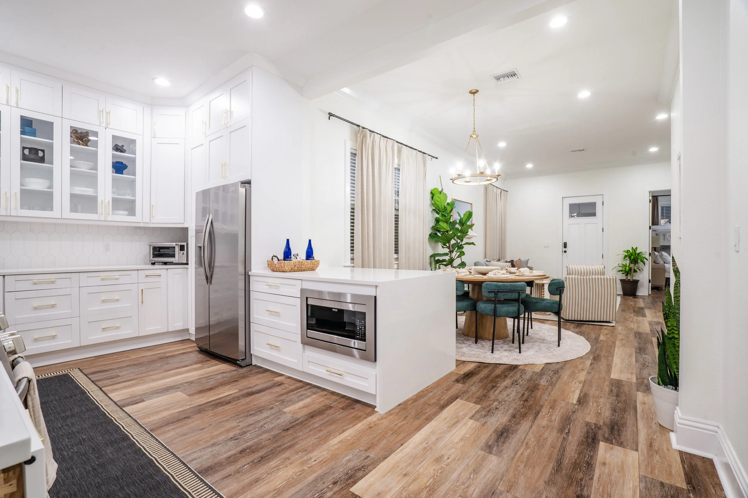 Open-concept kitchen and dining area with white cabinets, stainless steel appliances, wooden flooring, a round dining table with teal chairs, and a sitting area with sofa and striped accent chair, decorated with indoor plants and chandelier lighting.