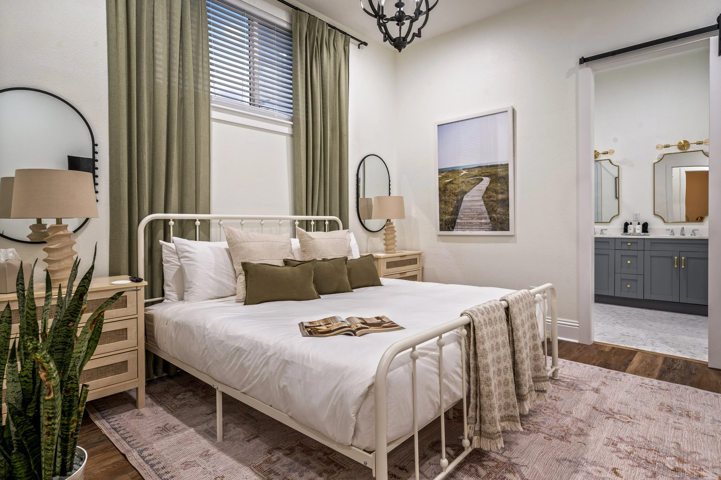 A bedroom with a white metal bed frame, beige and olive green pillows, a printed blanket, and a bedside table with a beige lamp, a mirror on the wall, a large framed landscape picture, and a bathroom visible through a door with a mirror and vanity.