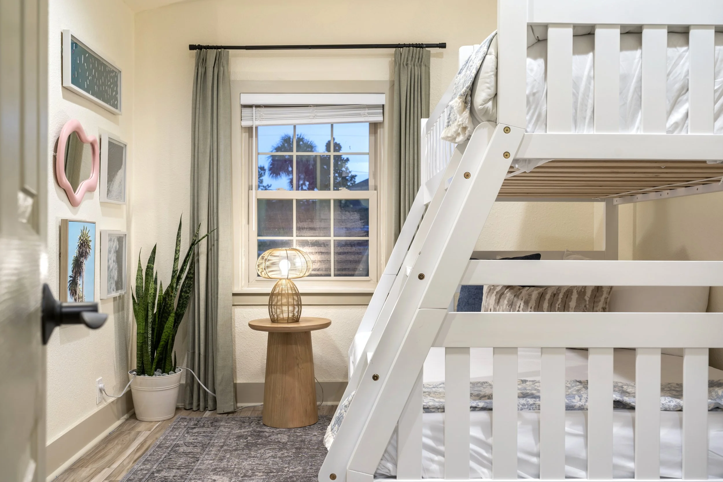 Bedroom with a bunk bed, window with curtains, lamp, framed pictures on the wall, and a potted plant.