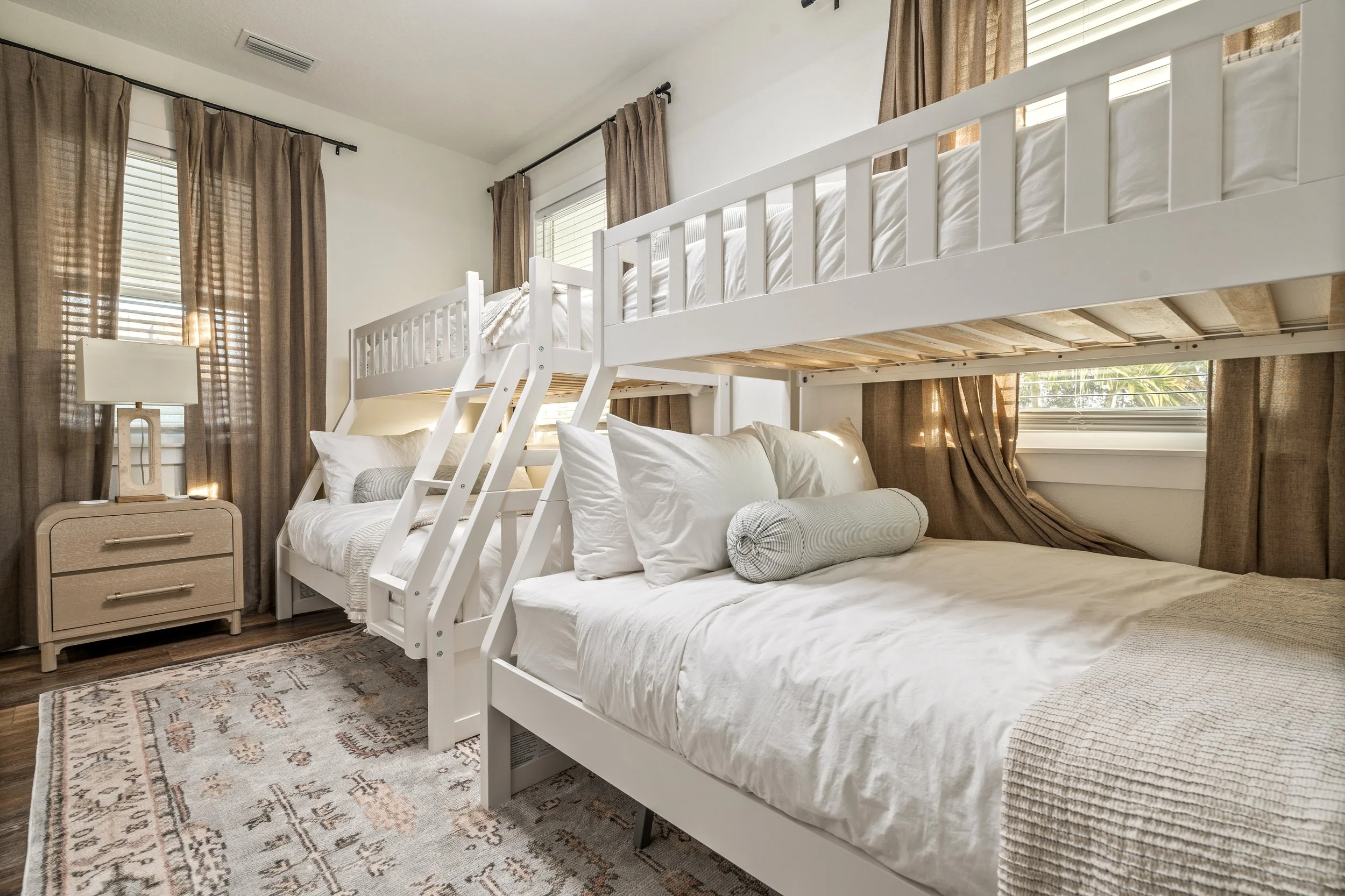 A bedroom with two white beds, one bunk bed with a ladder, beige curtains, a nightstand with a lamp, and a patterned rug.