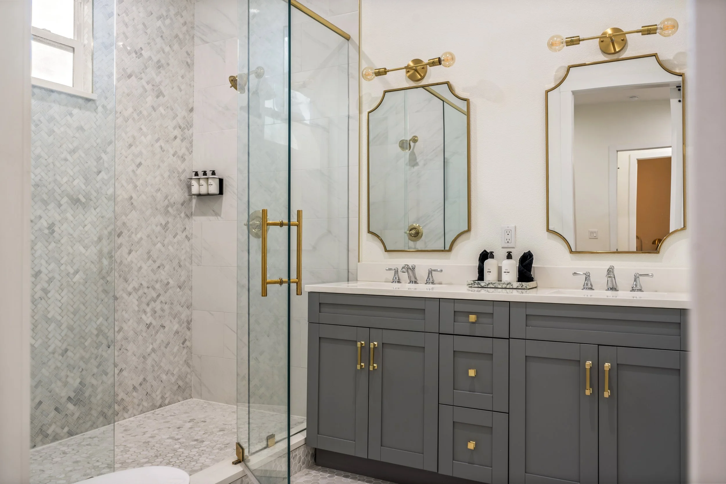 Modern bathroom with enclosed glass shower, gray vanity with gold handles, dual mirrors with gold trim, and two gold light fixtures with exposed bulbs.