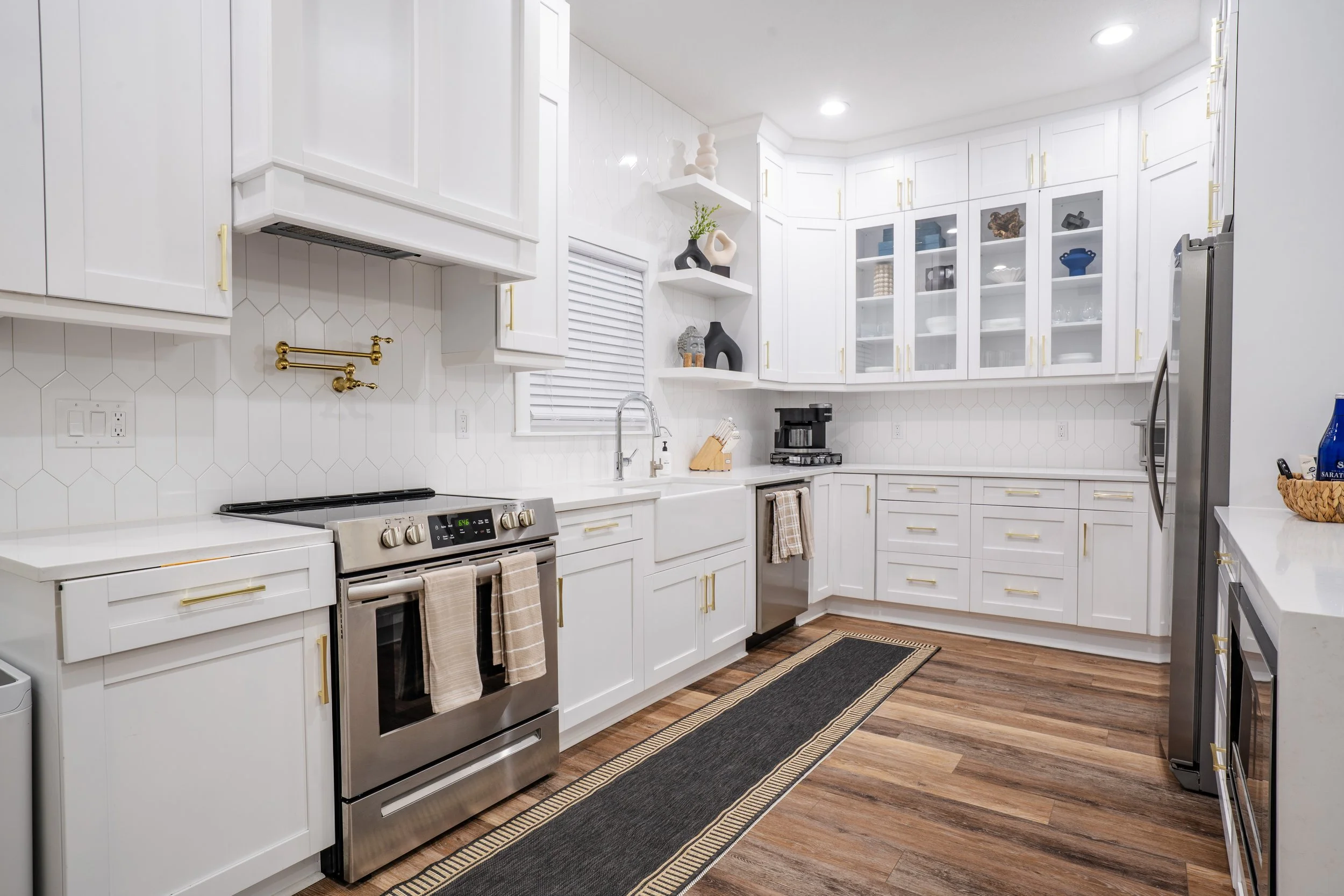 A modern kitchen with white cabinets, a stainless steel stove, a coffee maker, a refrigerator, and decorative vases on shelves. The kitchen has wooden flooring and a black rug.