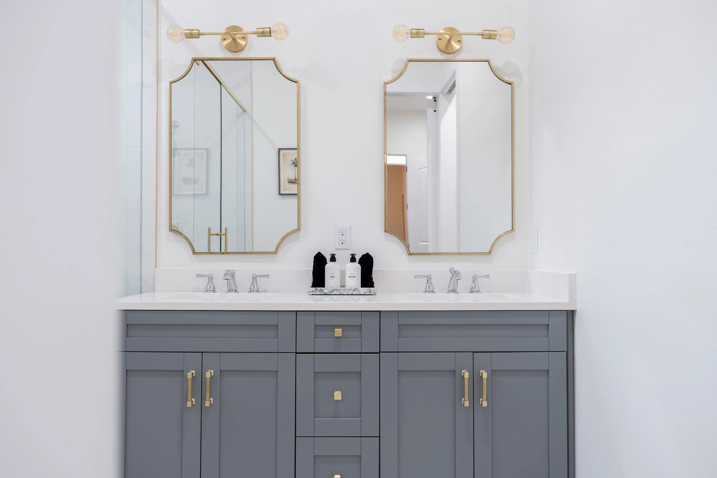 Bathroom vanity with a gray cabinet and a white countertop, two mirrors with gold frames, two wall-mounted lights, and soap and lotion bottles on a small marble tray.