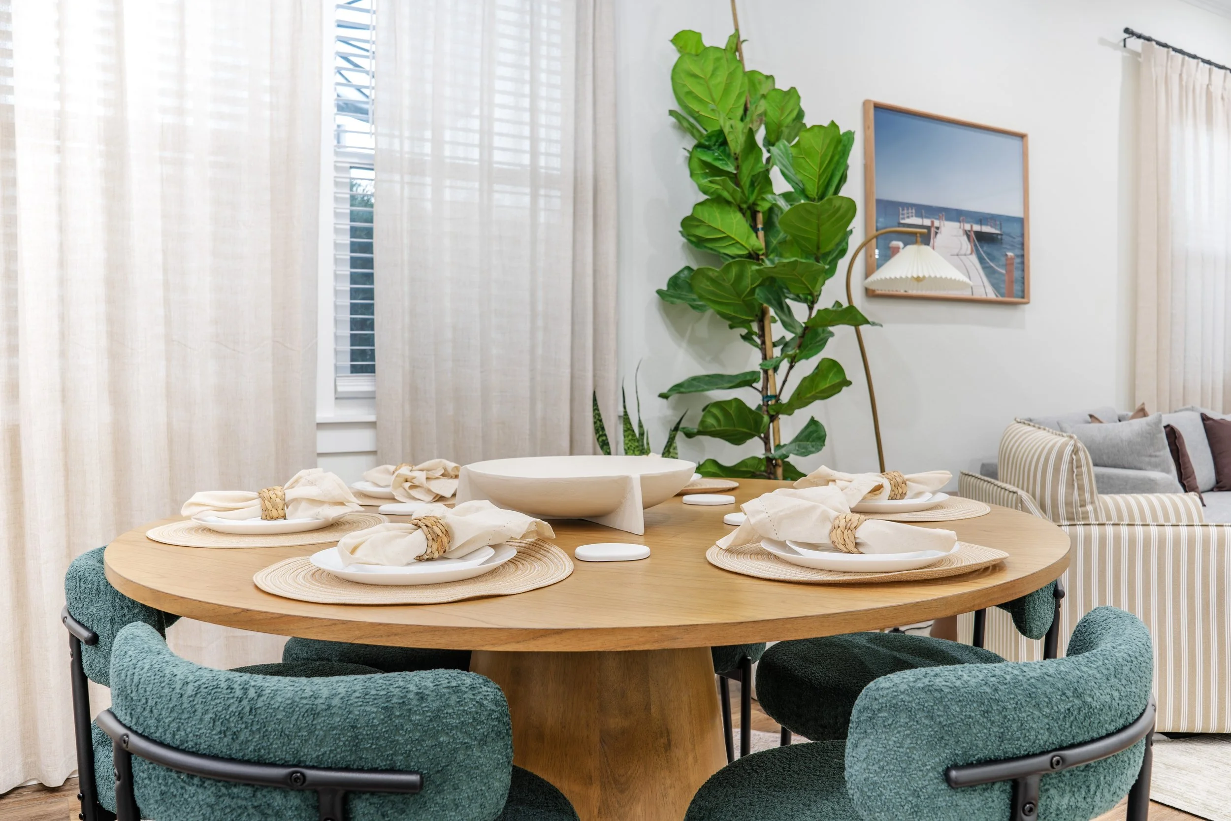 A round wooden dining table set with white plates, napkins, and woven napkin rings. Six green upholstered chairs surround the table. In the background, beige curtains cover windows, and a large potted plant with broad green leaves is next to a pictur