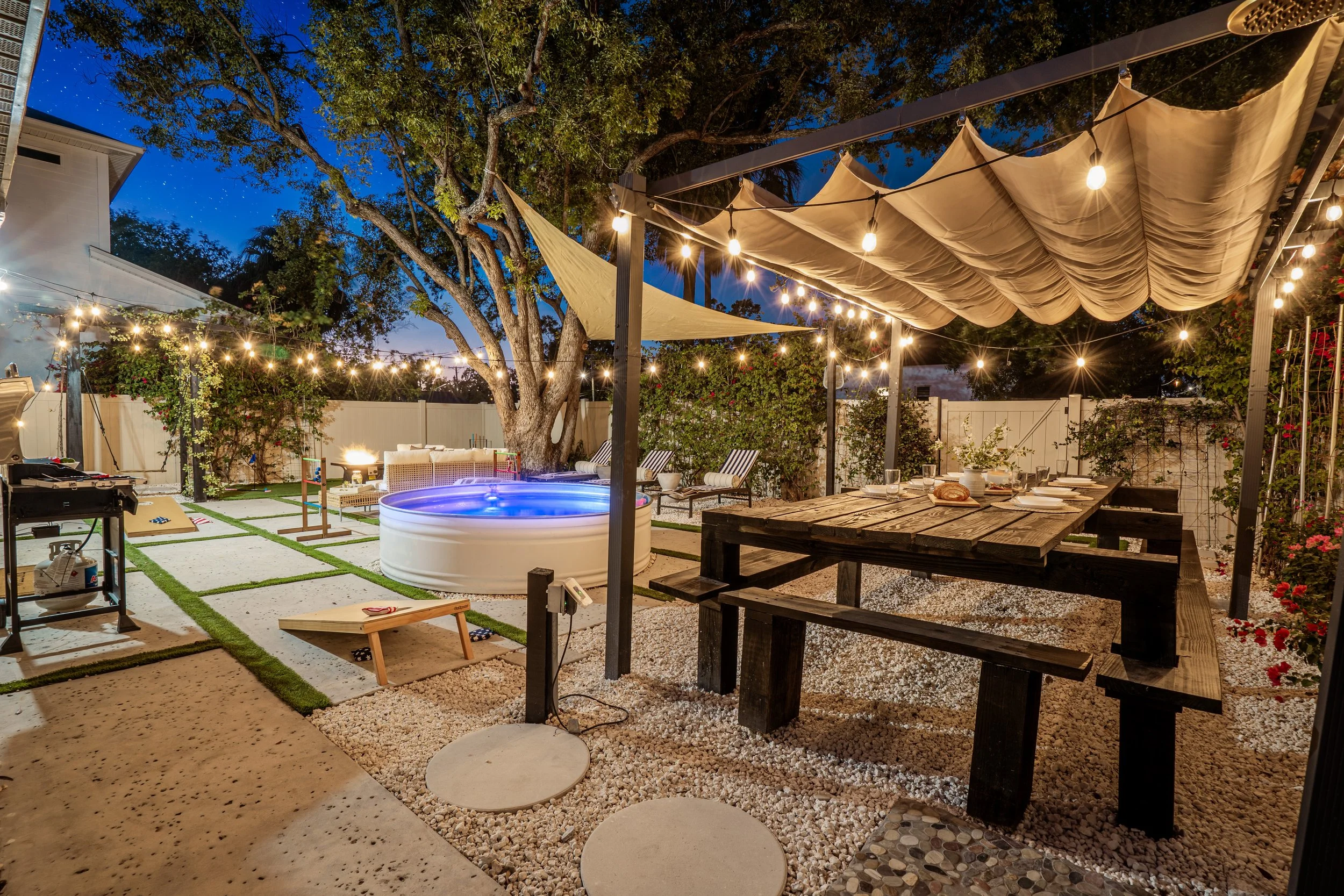 Nighttime backyard scene with string lights hanging over a dining table, a hot tub, a large tree, and outdoor seating, enclosed by a white fence.