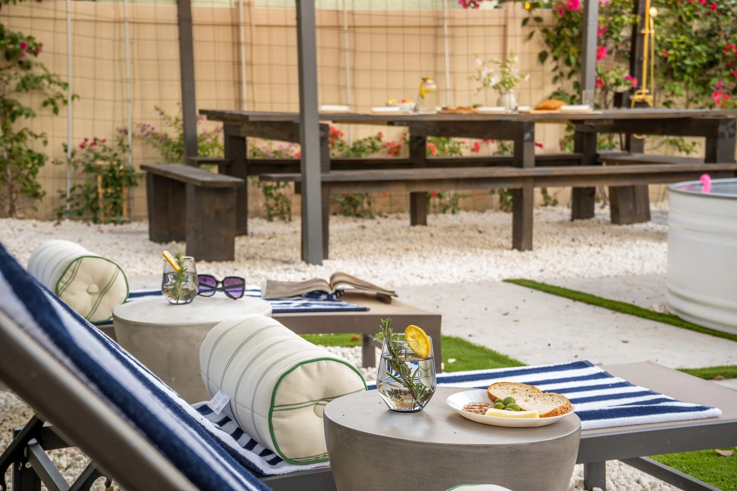 Outdoor patio scene with a table set for coffee and snacks, featuring a glass of water with a lemon slice, a plate with bread, cheese, and olives, and a cushioned lounge chair with striped fabric in the foreground. In the background, a wooden picnic 