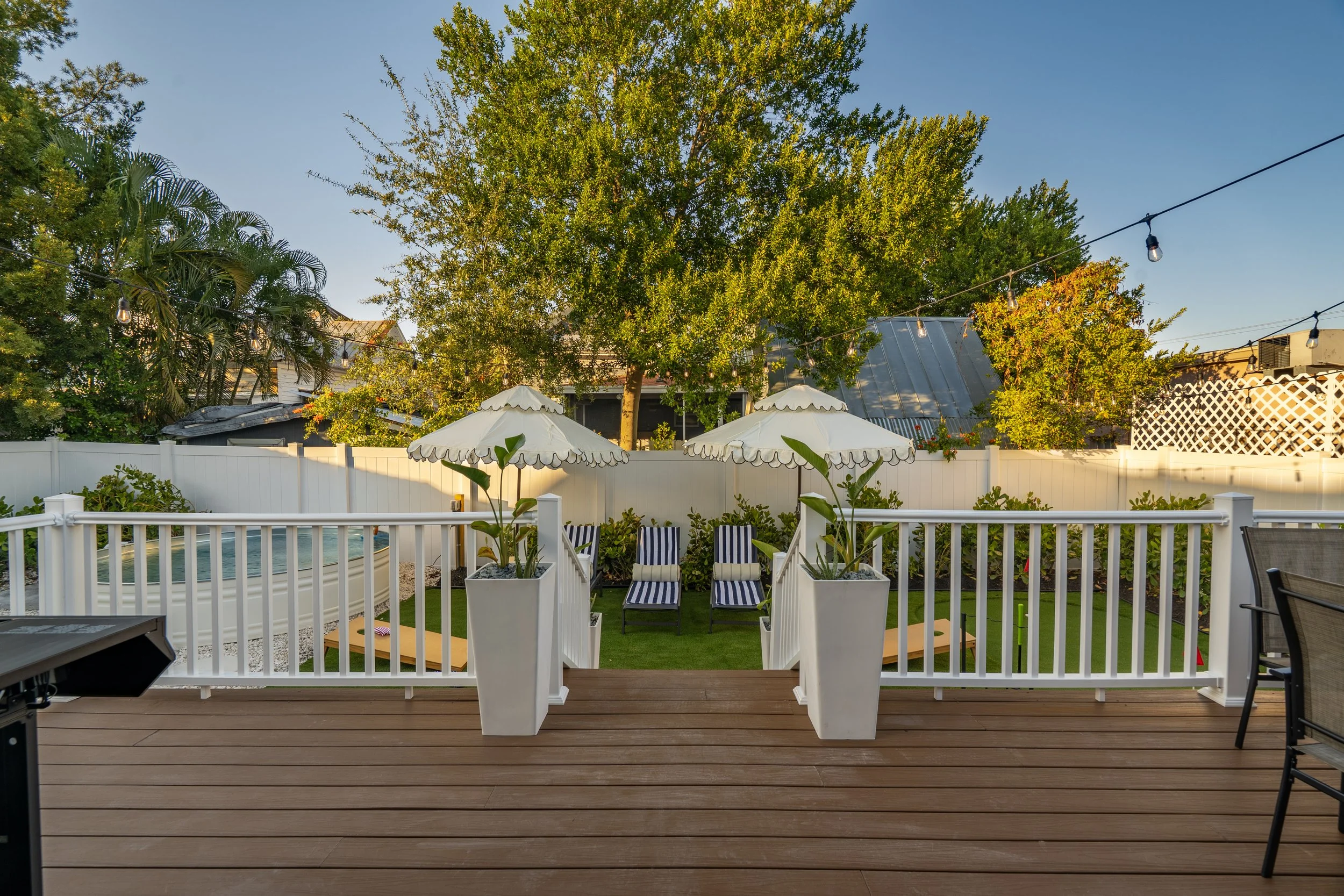 A backyard patio with a wooden deck, white fence, outdoor seating with striped chairs and umbrellas, trees, and string lights.