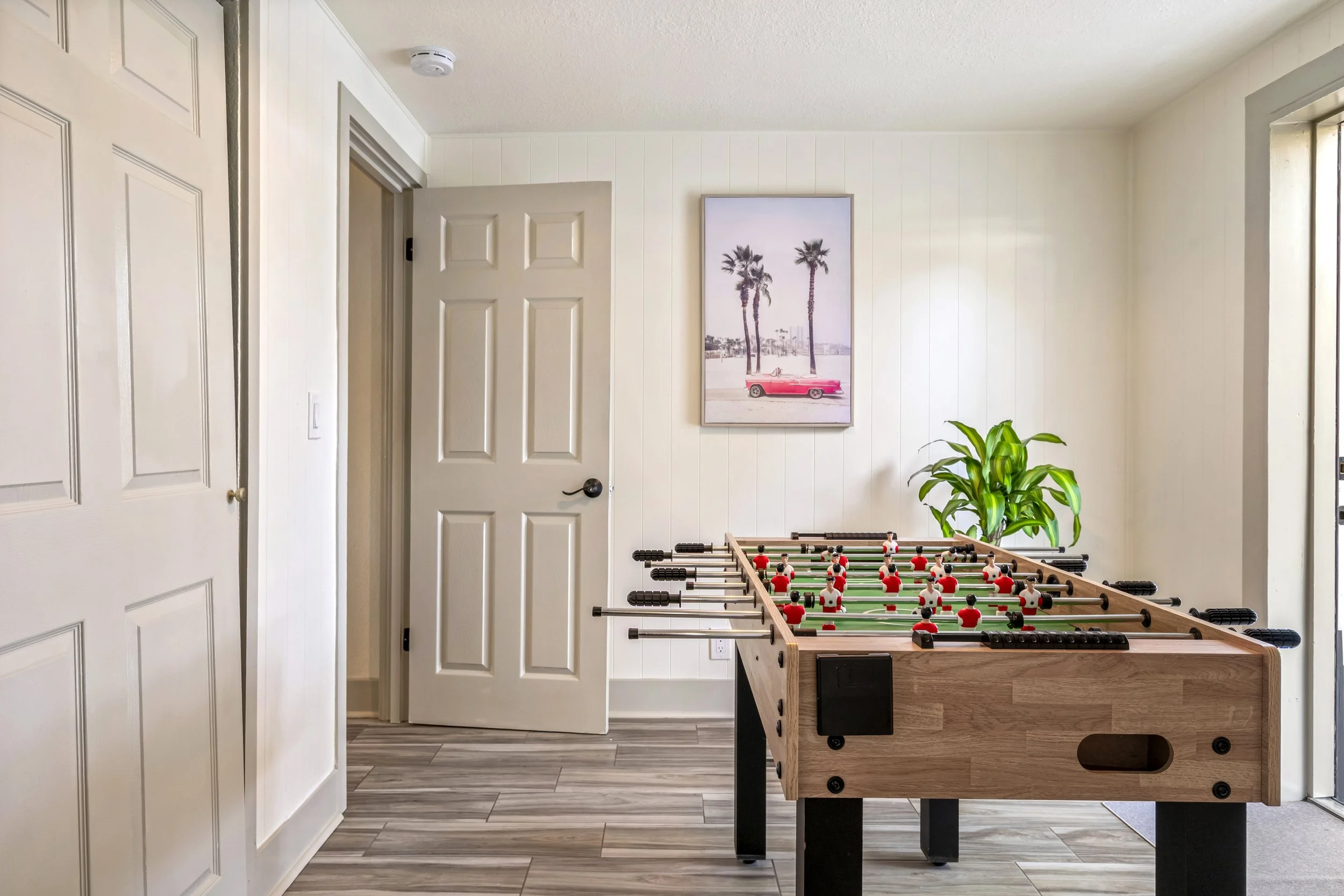 Interior of a room with white paneled walls, an open door, a framed picture of palm trees and a pink car, a green plant, and a foosball table.