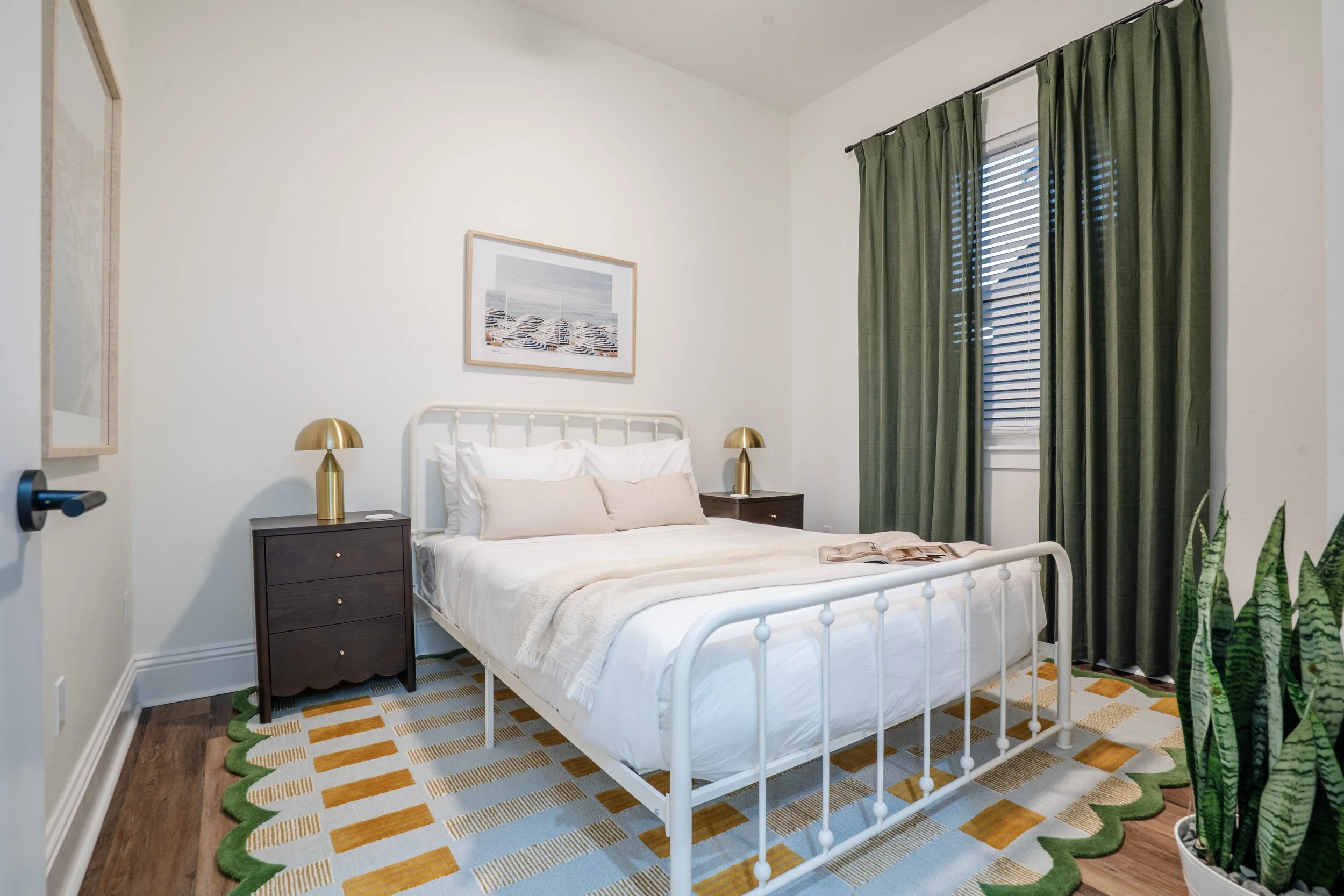 A bedroom with a white metal bed frame, white bedding, two bedside tables with gold lamps, a colorful patterned rug, green curtains, a window with blinds, and a wall art picture of beach chairs and umbrellas.