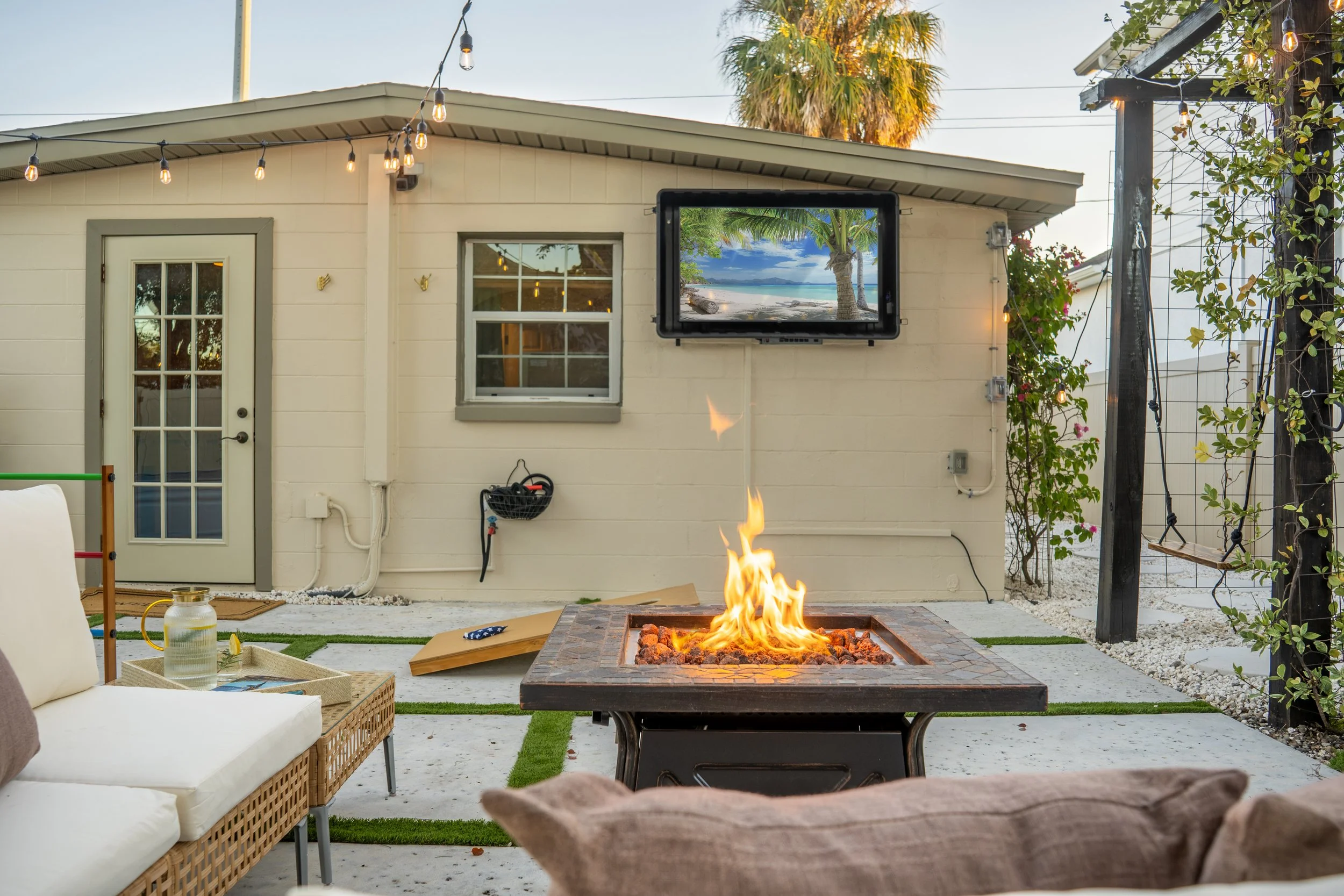Outdoor patio scene with a fire pit, seating, string lights, a wall-mounted TV displaying a beach scene, and a background with palm trees and a clear sky.