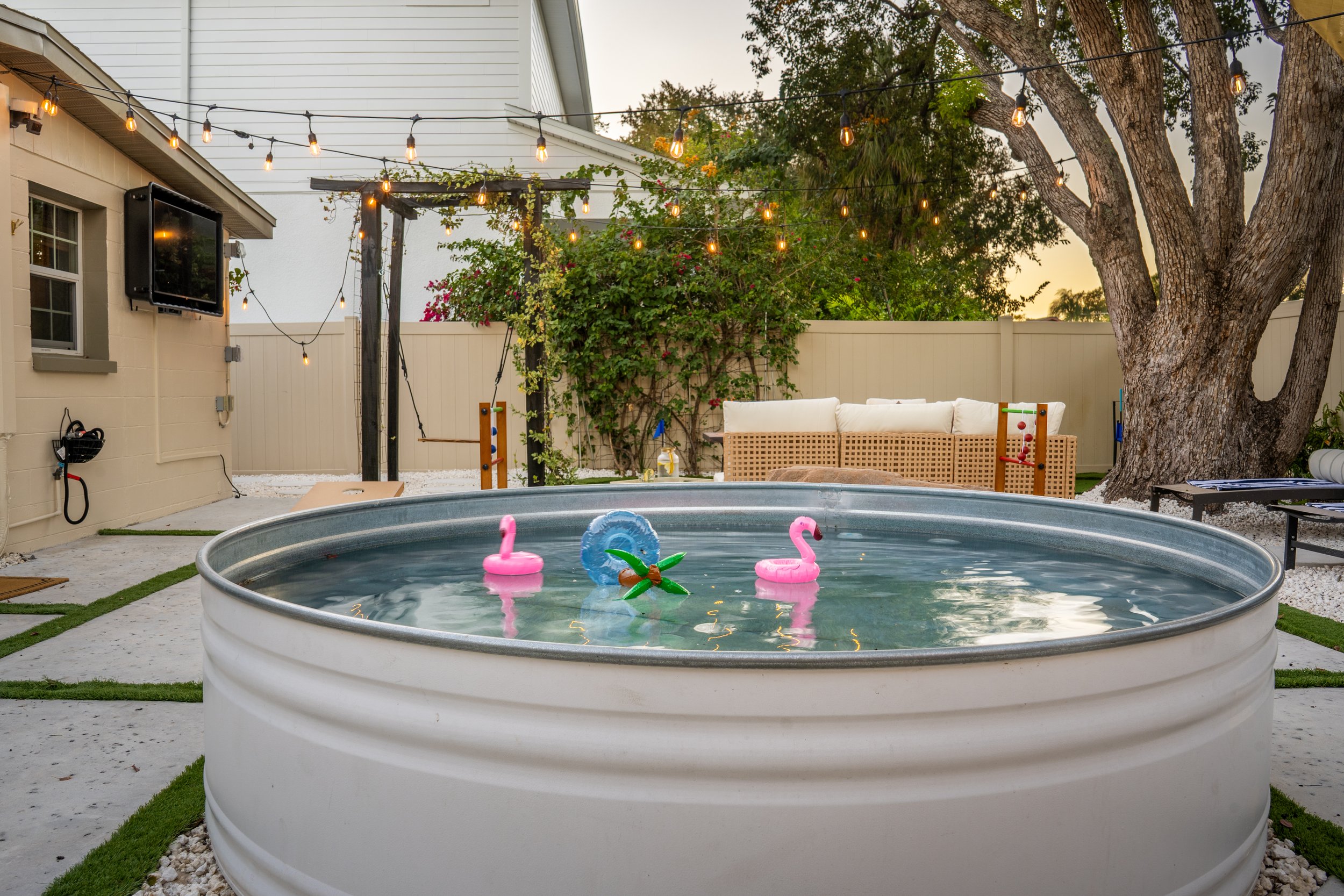 Backyard patio with string lights hanging above a small inflatable pool with floating flamingo and pineapple toys, a white sofa, a tree, a fence, and adjacent house wall with a wall-mounted TV.