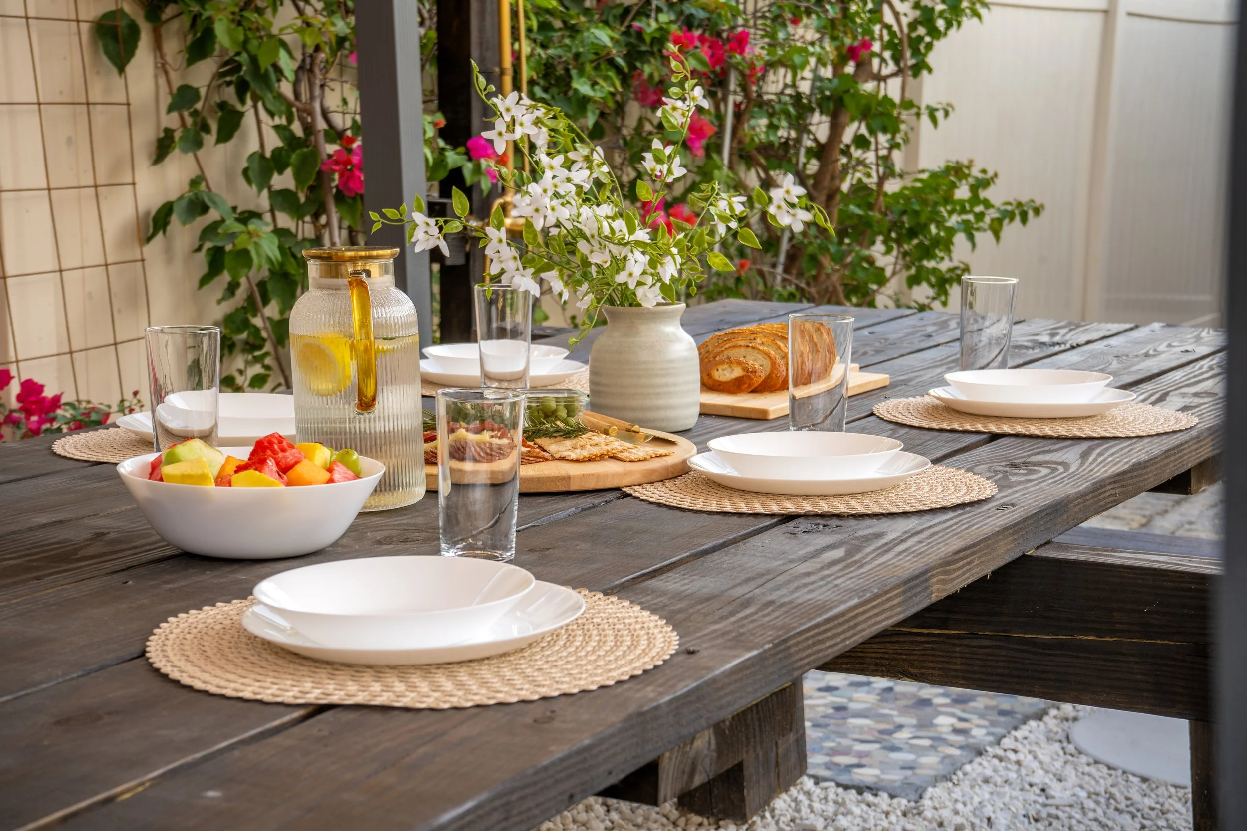 A rustic outdoor dining table set with white plates on woven placemats, clear glasses, a pitcher of lemonade, a bowl of fruit salad, sliced bread, and a vase with white flowers. In the background, there are blooming pink and red bougainvillaea plants