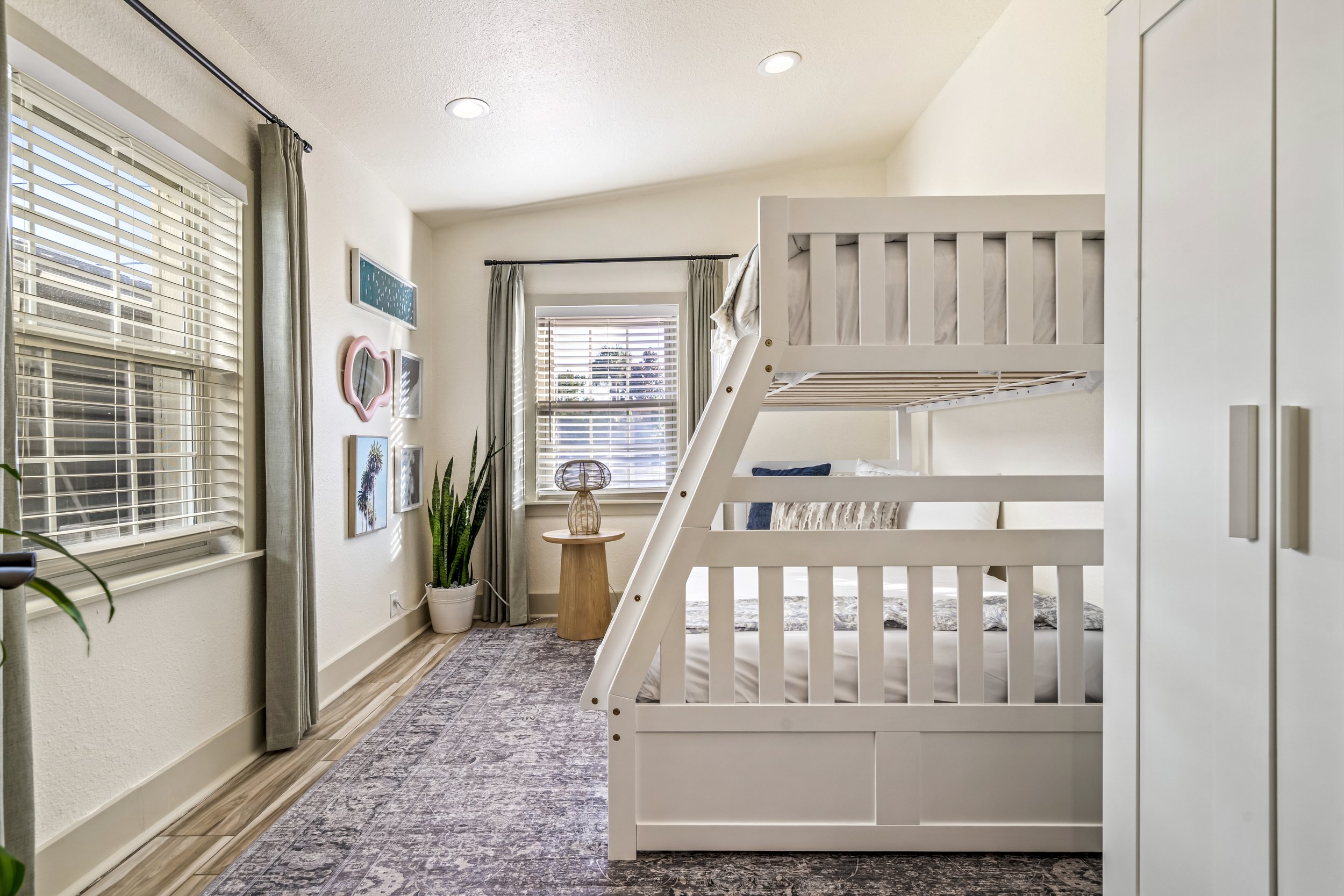 A bedroom with large windows, light-colored walls, and a white bunk bed. There are curtains on the windows, a small wooden side table with a lamp, and artwork on the wall. The room has a cozy and modern decor with a patterned rug on the floor.