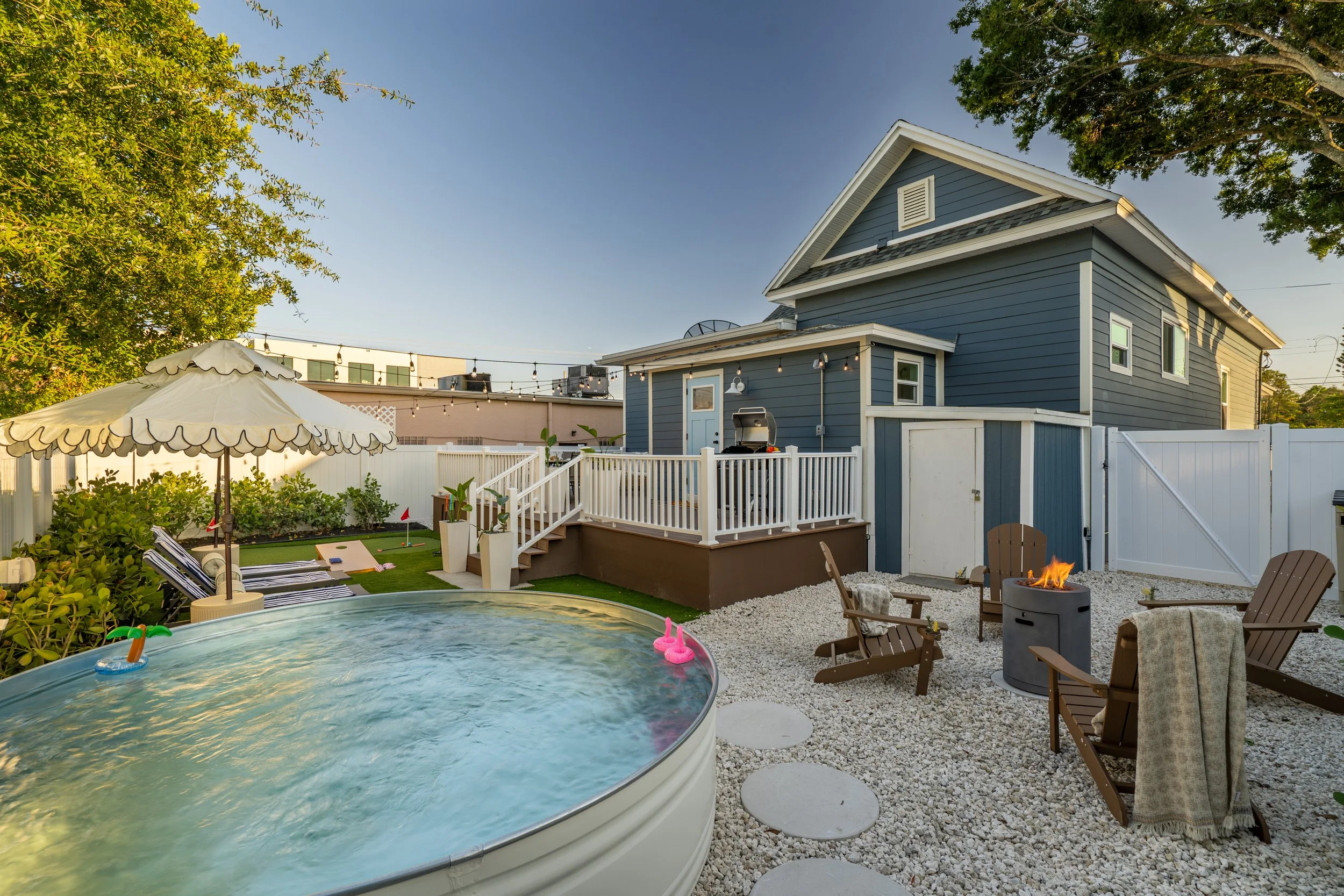 A backyard with a swimming pool, lounge chairs, a patio with an outdoor fire pit, a blue house with a white fence, and a large tree, all illuminated by evening sunlight.