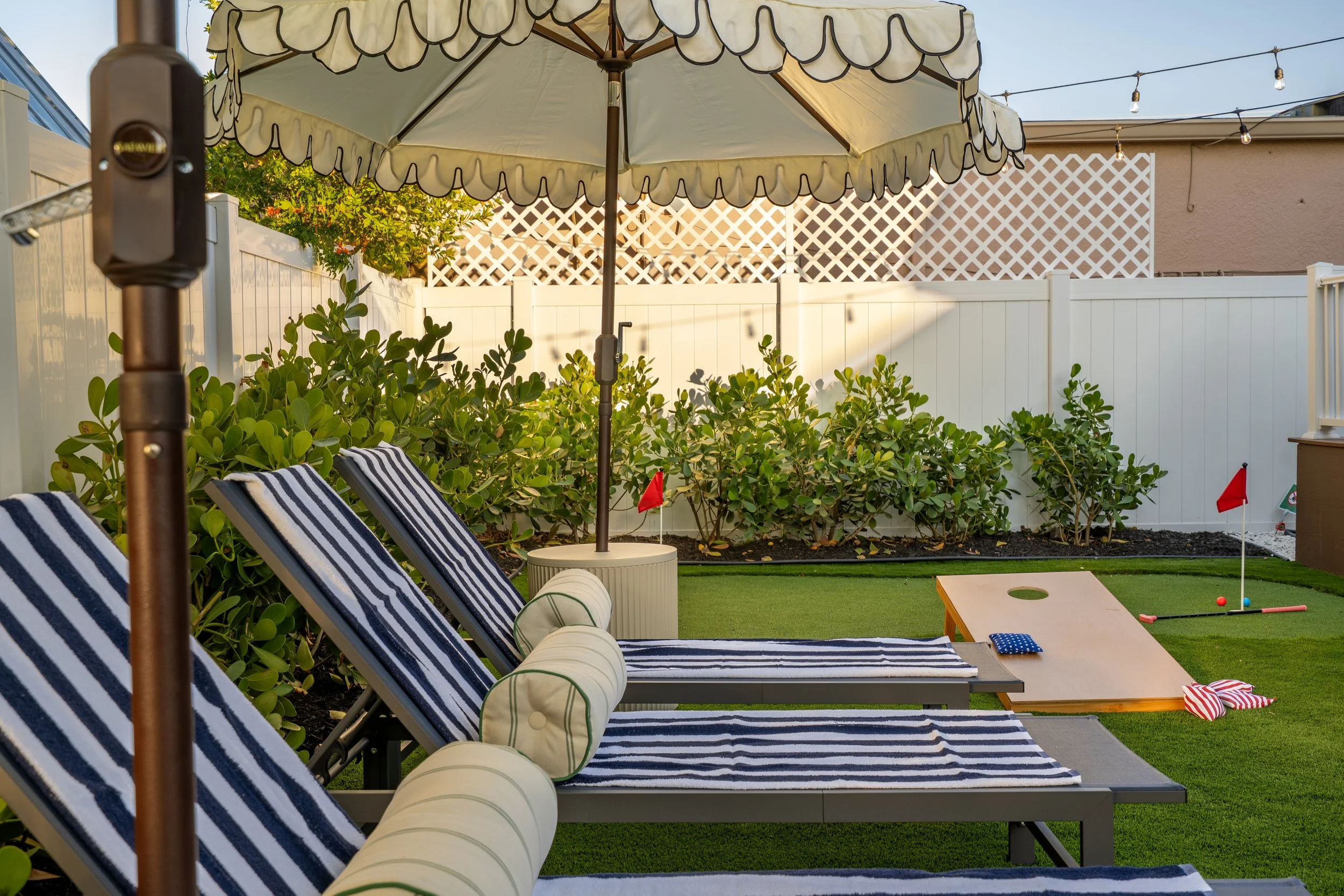 A backyard scene with a large beige umbrella, striped blue and white lounge chairs, green plants, a white fence, a small putting green with miniature golf flags, and a string of outdoor lights.