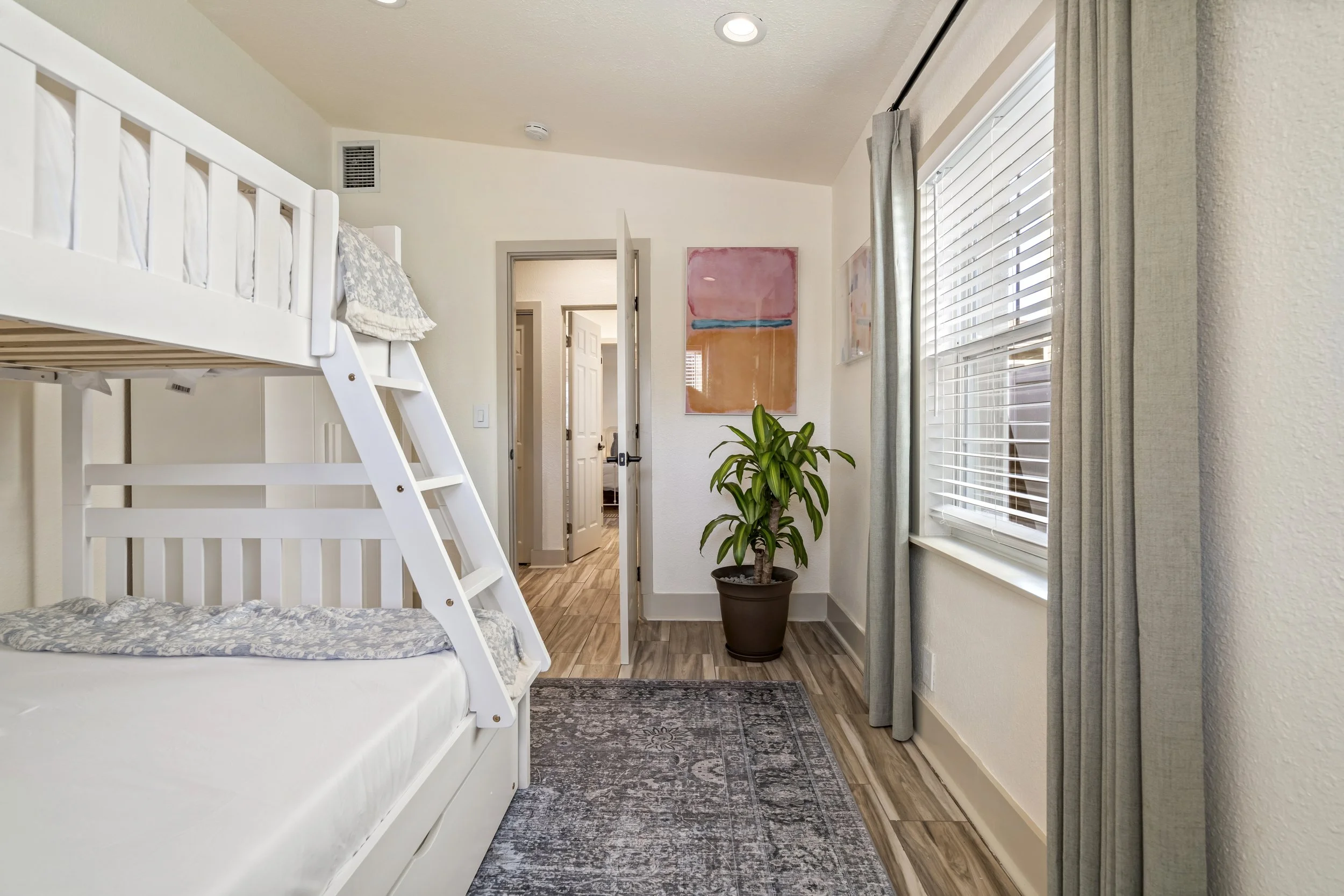 A bedroom with white walls and a ceiling, featuring a white bunk bed with gray and white bedding. There are large windows with white blinds and gray curtains, a potted green plant, and colorful abstract artwork on the wall. The floor has wood planks 