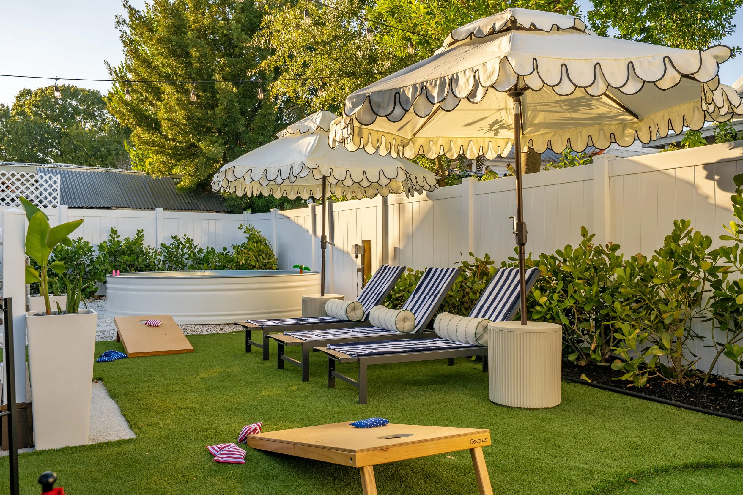A backyard area with three white patio umbrellas, striped lounge chairs, a white fence, green bushes, potted plants, string lights, and a cornhole game on a grassy lawn.