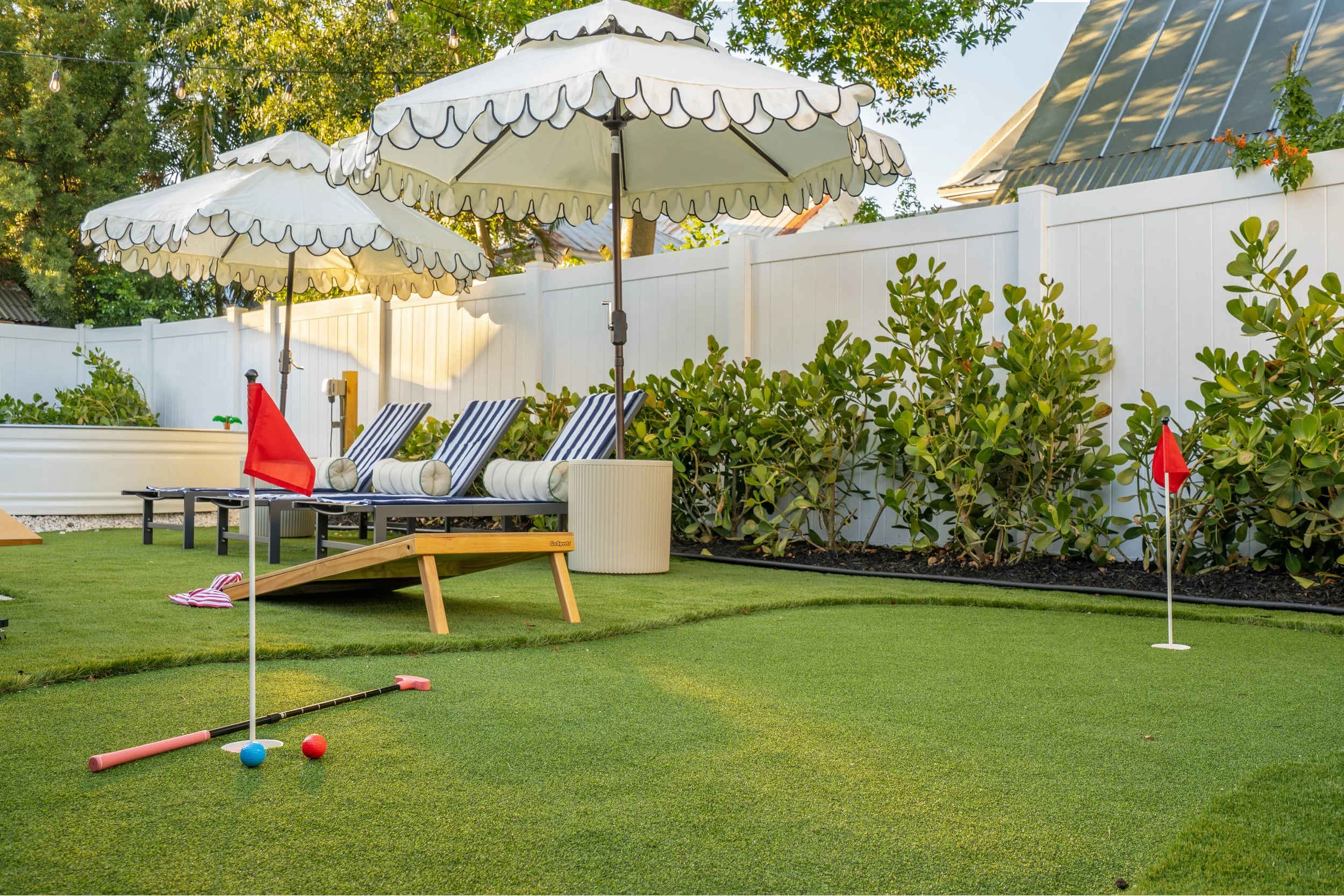 Backyard with three large cream-colored patio umbrellas, lounge chairs with striped cushions, a white fence, and a putting green with golf balls and golf club, surrounded by green shrubs and trees.