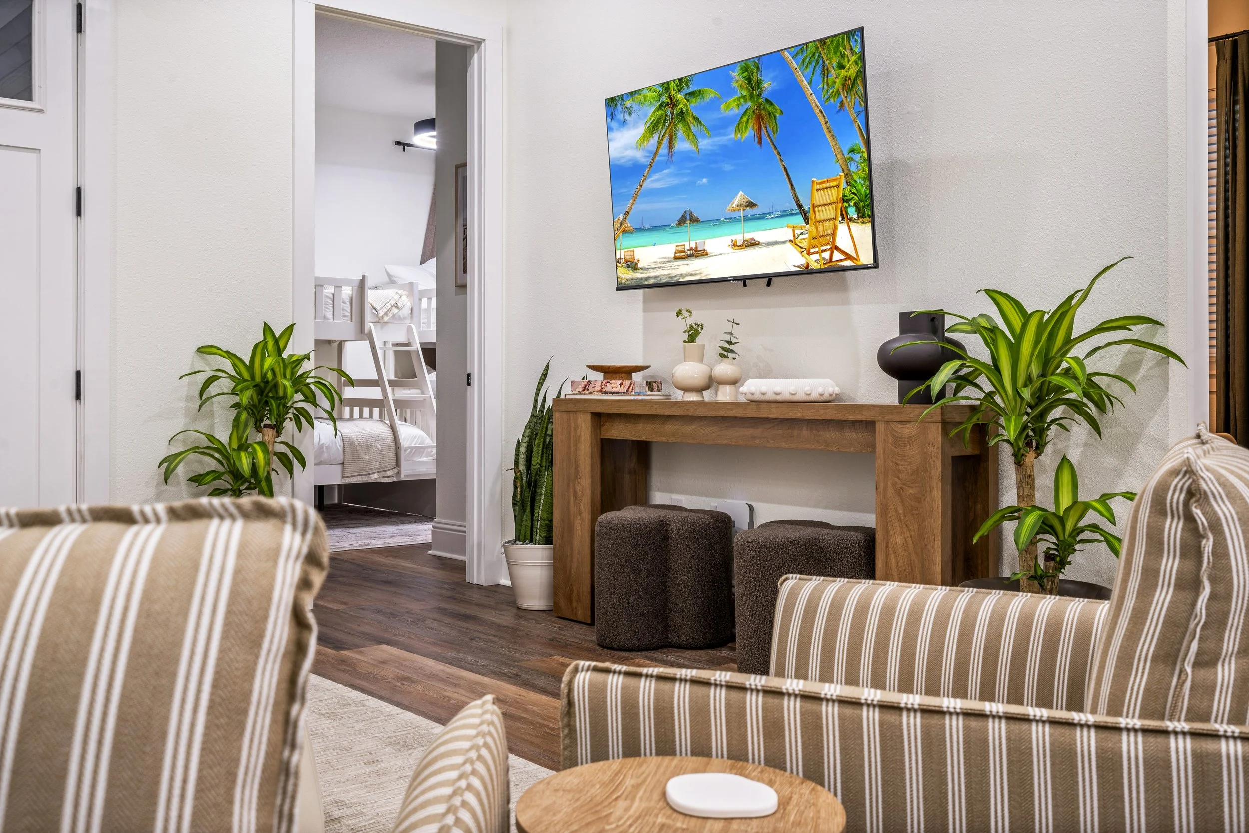 Living room with striped beige and white chairs, potted plants, a wooden console table with decorative items, a television displaying a beach scene, and a doorway leading to a bedroom with a white bunk bed.