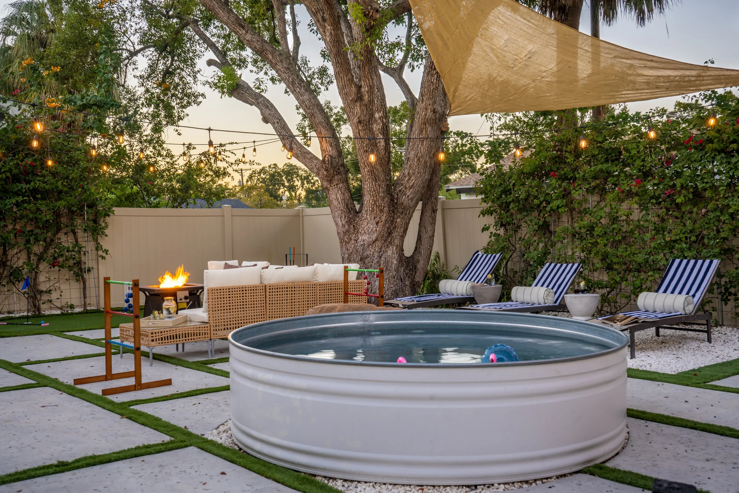 A backyard patio with string lights hanging above, a large tree at the center, a white fence, outdoor furniture including a white sofa with pillows, striped lounge chairs, a small fire pit, and an above ground plastic pool filled with water.