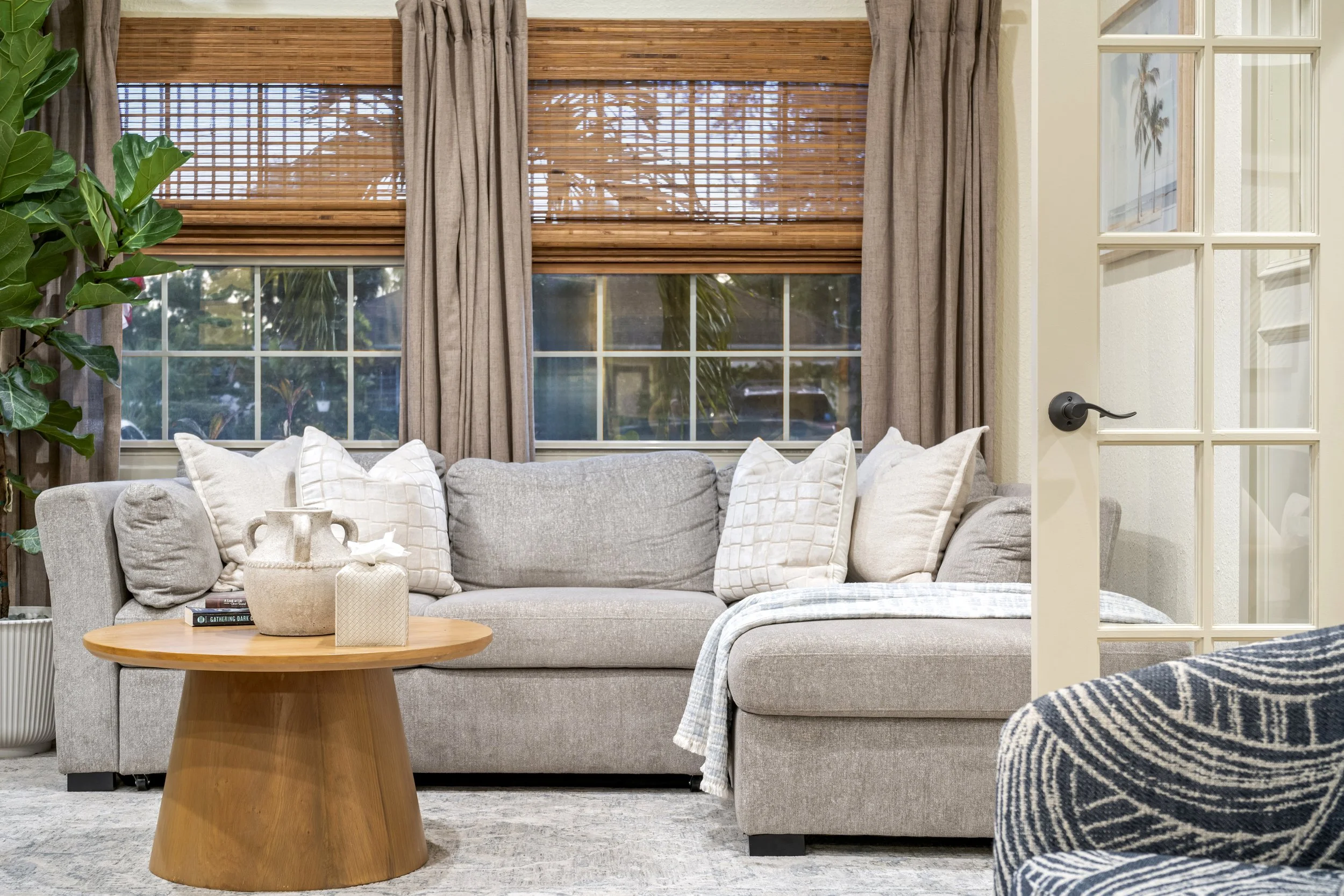 Living room with a beige sofa, white pillows, a round wooden coffee table with decorative items, large windows with bamboo shades and beige curtains, a tall potted plant, and a partial view of a black and white striped armchair.