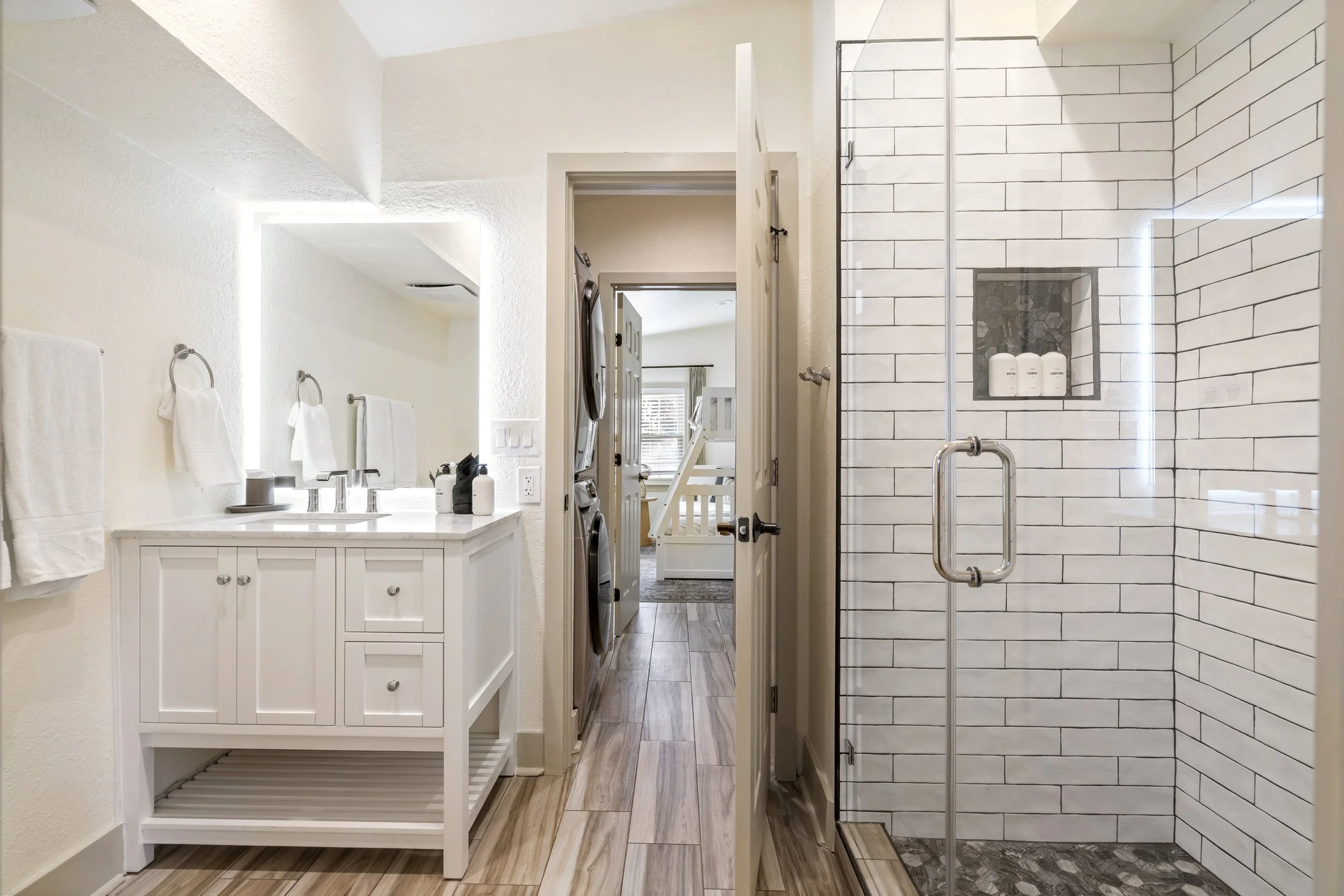 Bathroom with a white vanity, towel racks, and a walk-in shower with white subway tiles and bottles of shampoo and soap.