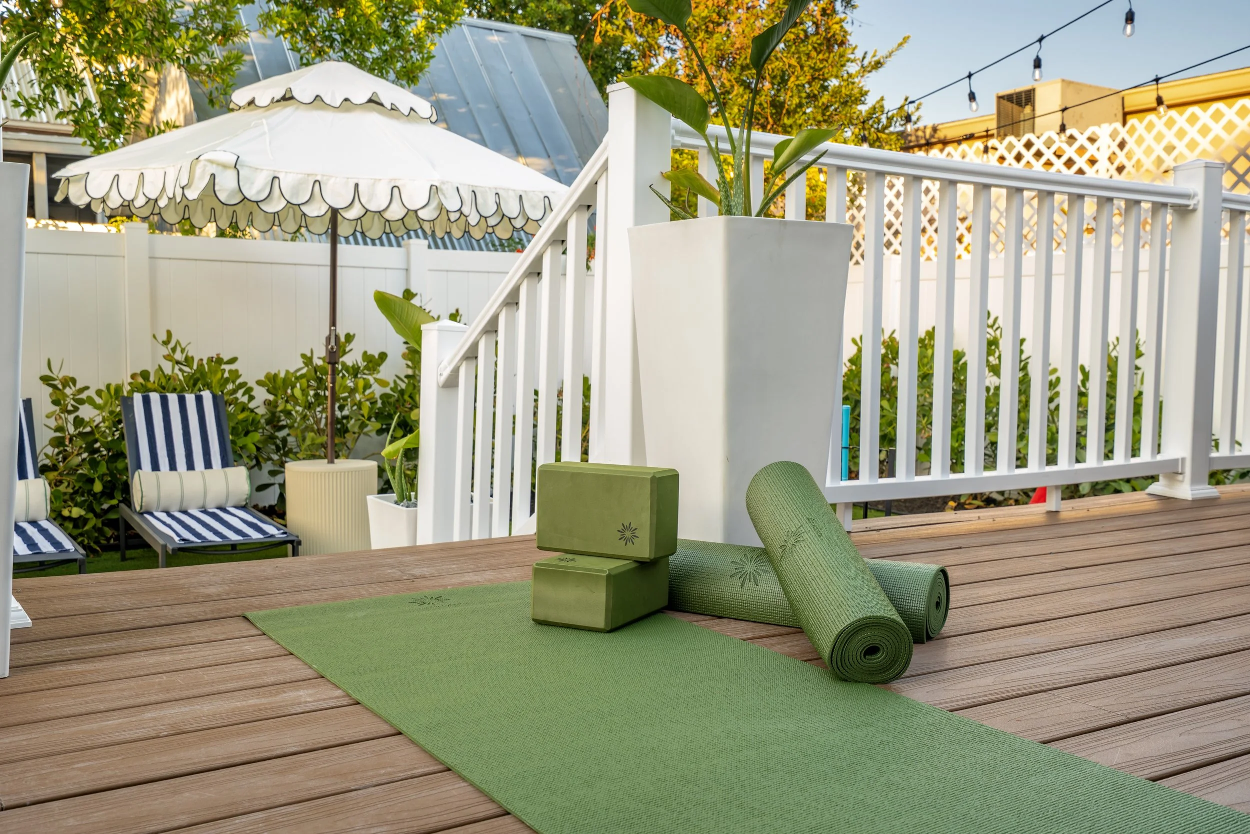 A backyard patio featuring lounge chairs with striped cushions, a white fence, potted plants, a green yoga mat with a rolled-up yoga mat on top, and outdoor umbrellas with scalloped edges, surrounded by trees and string lights.