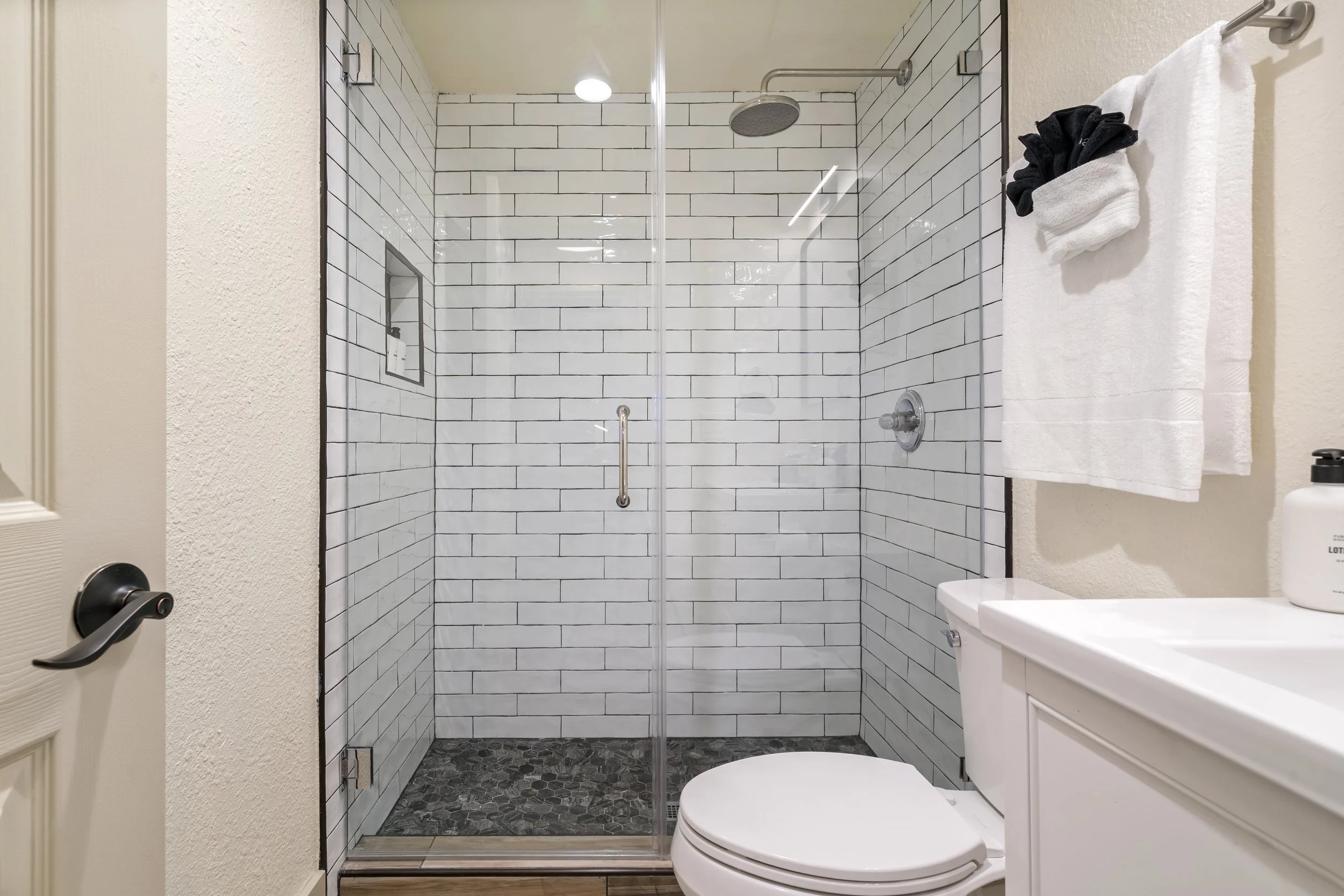 A modern bathroom shower area with white subway tiles, a round overhead shower, and a clear glass door. There are white towels hanging on a silver towel rack on the right and a white toilet below. The left side shows a textured wall and part of a doo