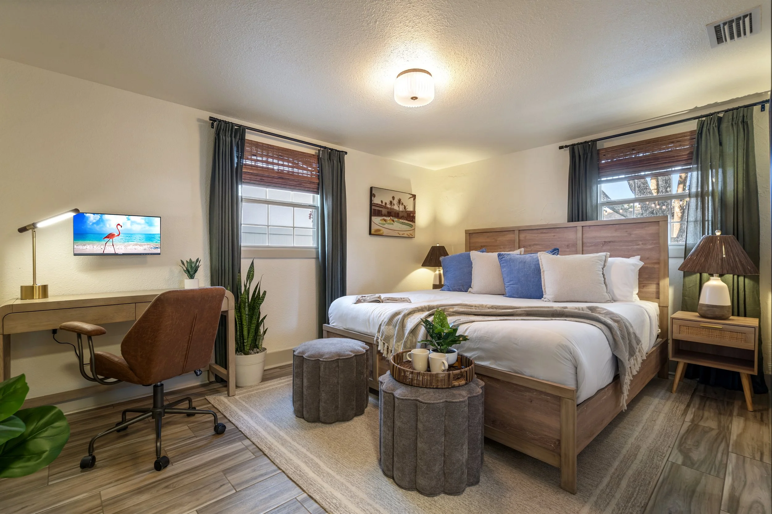 A cozy bedroom with a large bed featuring white and blue pillows, wooden headboard, and side tables with lamps. Two windows with dark curtains and wooden blinds let in natural light. There are houseplants, a small tufted ottoman, and a tray with coff
