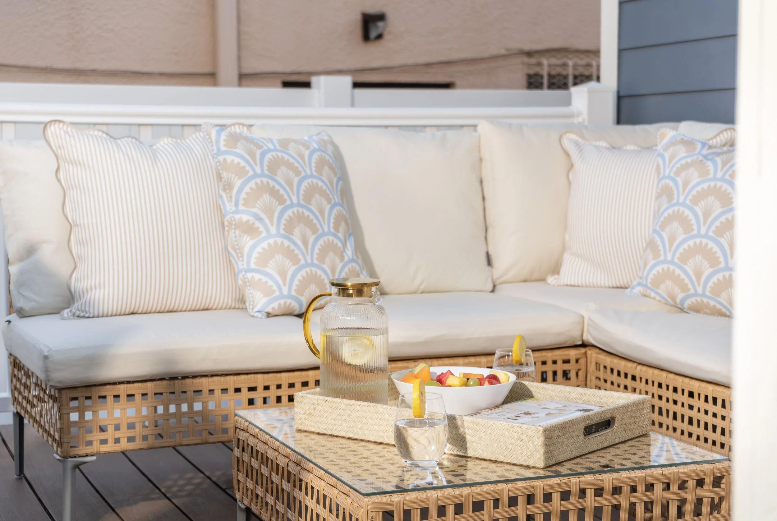 Outdoor patio seating area with a rattan sofa decorated with light-colored and patterned pillows, a glass pitcher of water with lemon, a bowl of fruit, a glass of water with lemon on a wicker table, and a tray with brochures or magazines. The setting