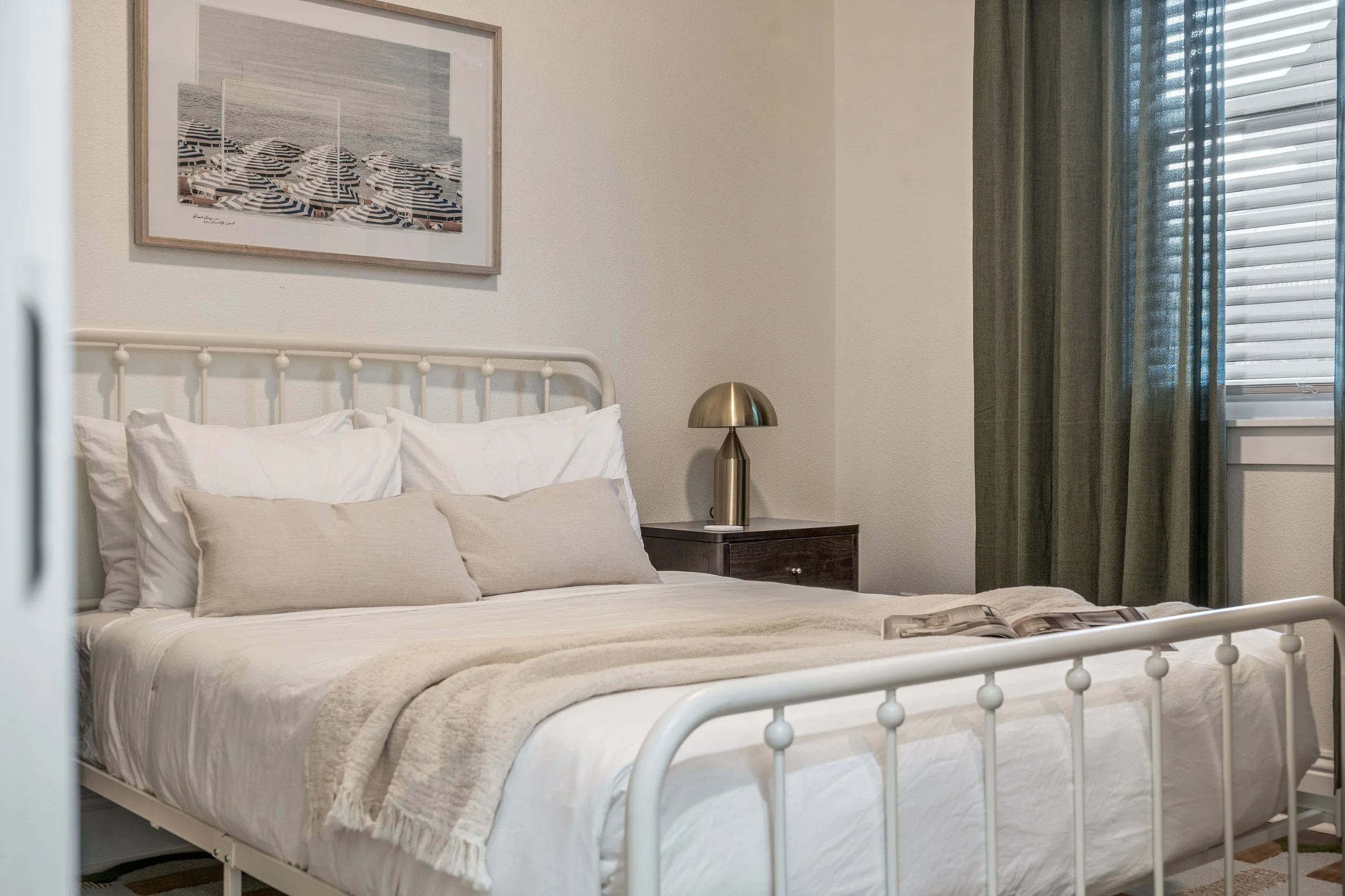 A cozy bedroom with a metal bed frame, white bedding, a beige blanket, and pillows. There is a framed picture of umbrellas on the wall, a gold bedside lamp, and a window with green curtains.