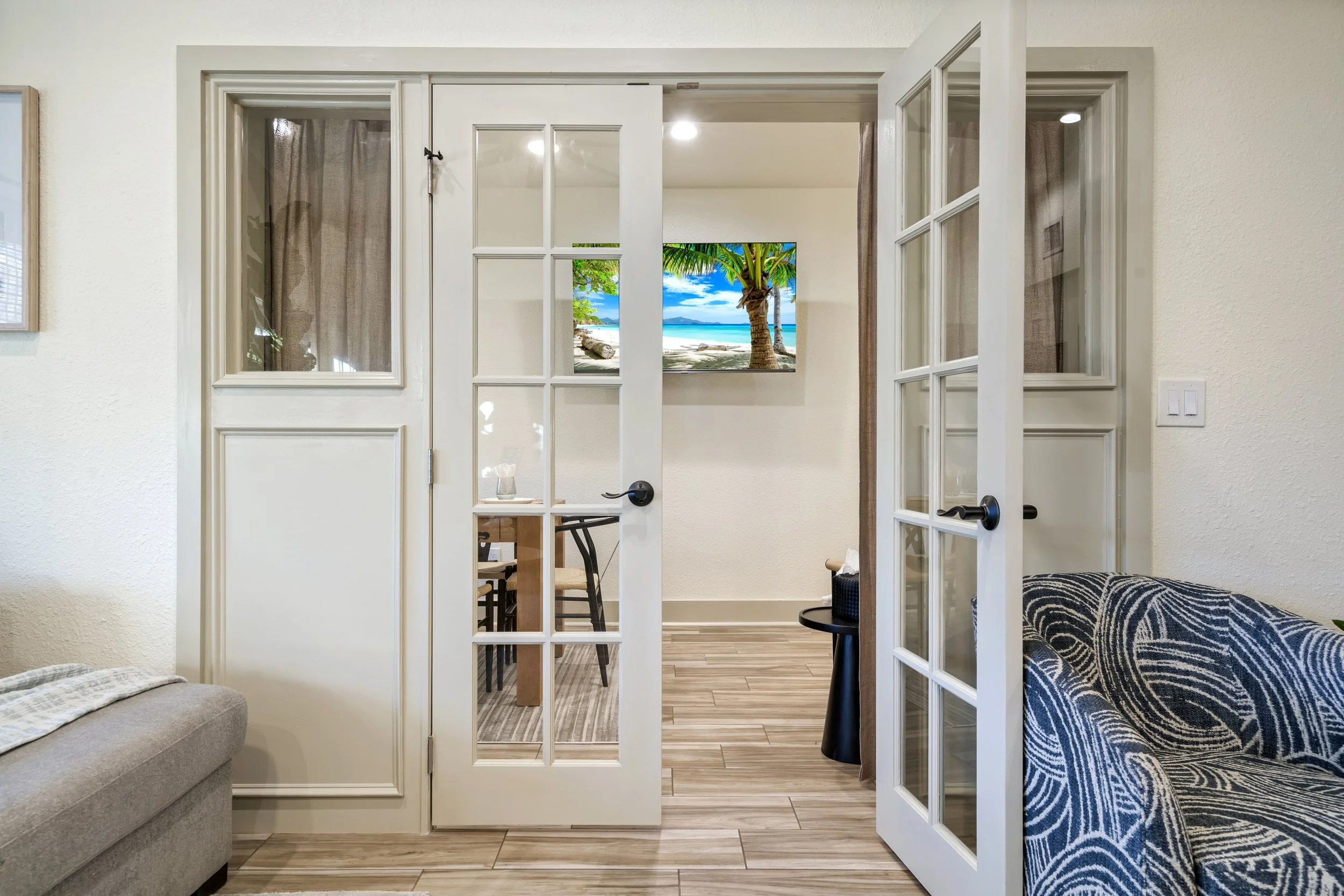 Living room with glass doors leading to a dining area, a wall-mounted TV showing a beach scene with palm trees, and a sofa with a patterned blanket.