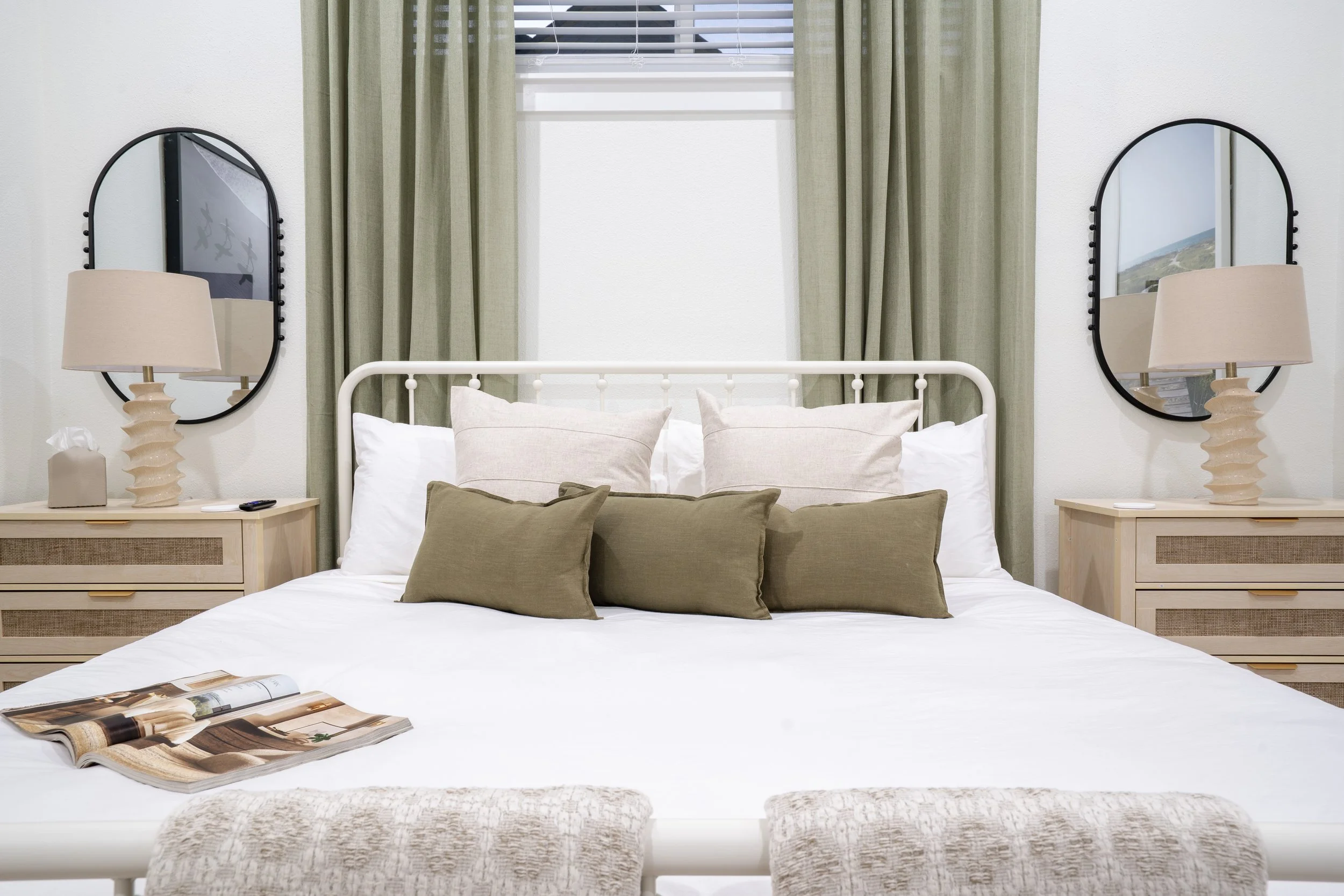 A neatly made bed with white sheets and multiple beige and green pillows. Two oak nightstands with beige lamps and a tissue box are on either side of the bed. There are two oval mirrors on the wall behind the bed, curtains and a window visible in the