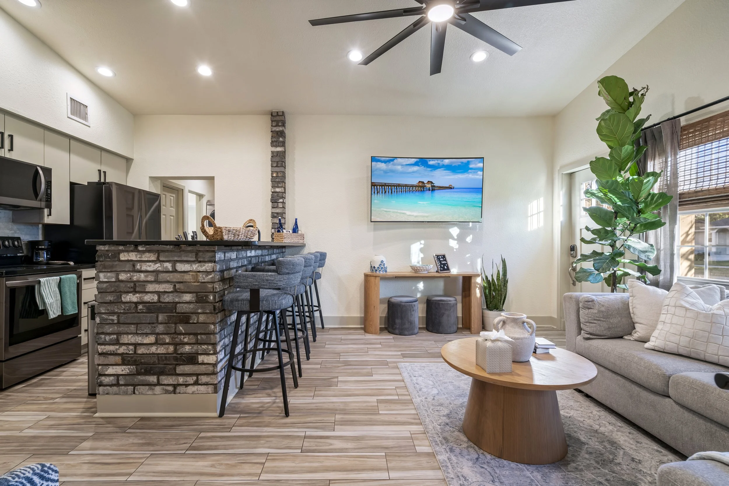 Open-concept living room and kitchen with brick island, bar stools, a wall-mounted TV showing a beach scene, a large green plant, and a ceiling fan.