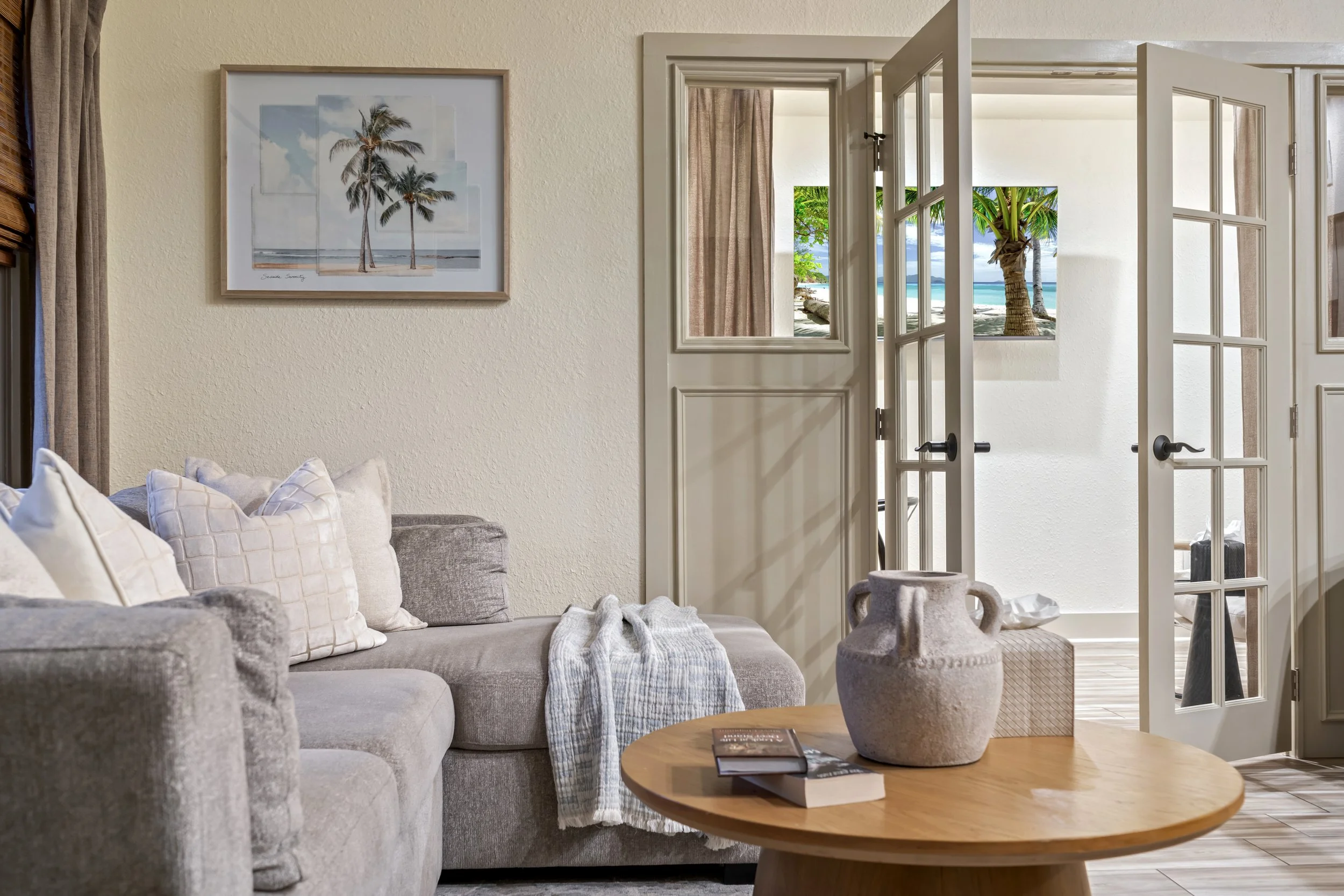 Living room with beige sofa, decorative pillows, wooden coffee table with books, ceramic vases, and view of tropical beach through open French doors.