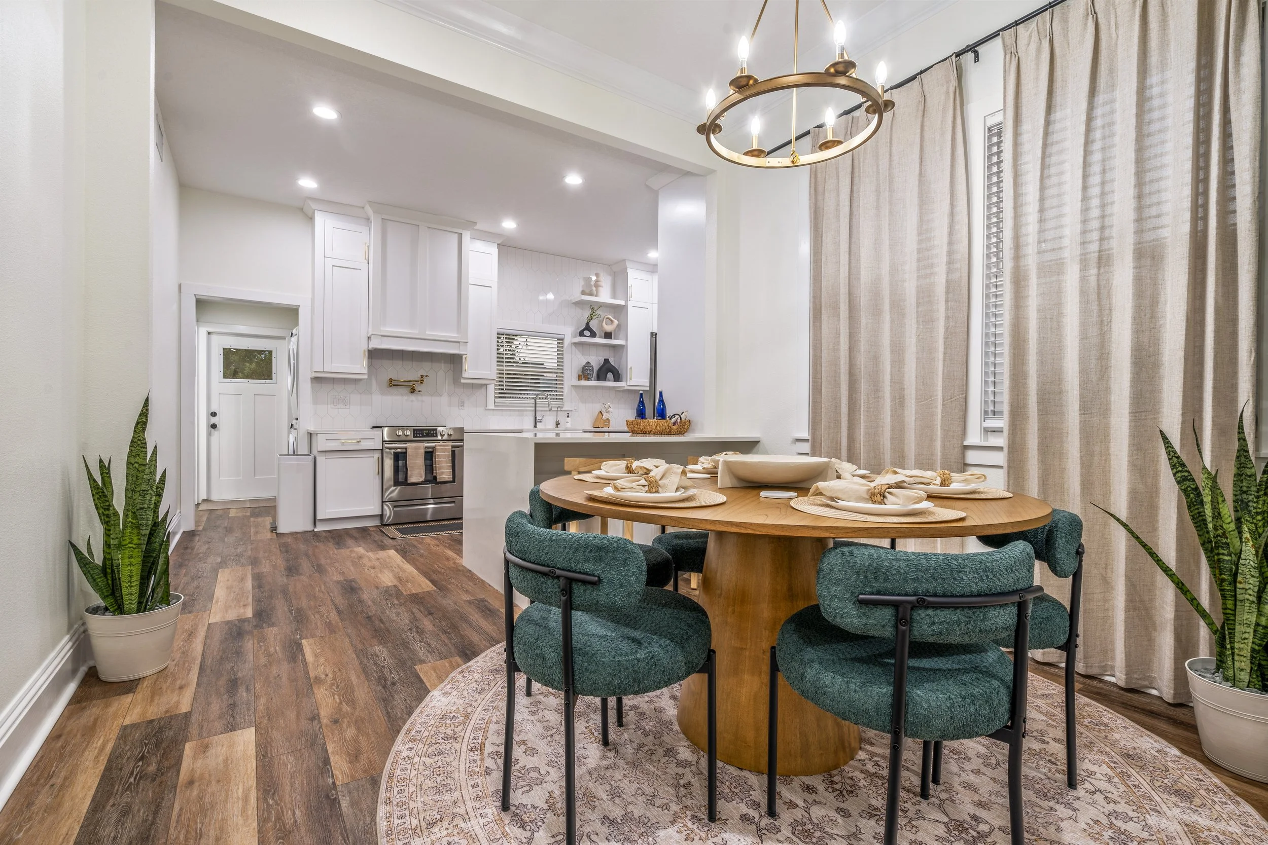 Dining area with a round wooden table set with cream-colored napkins and plates, surrounded by green upholstered chairs, positioned on an ornate rug, with large beige curtains, and plants in white pots nearby. In the background, a white kitchen with 