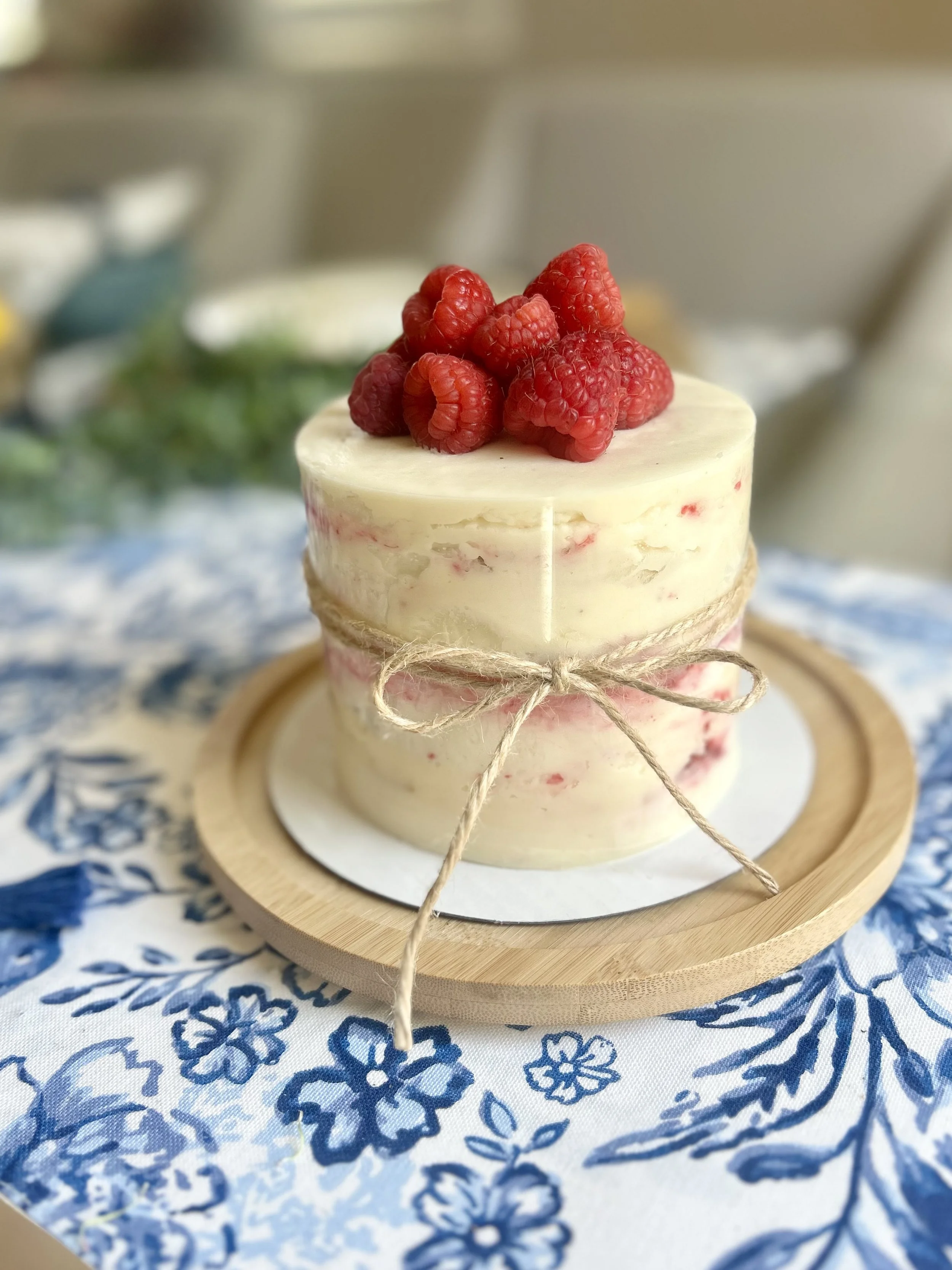 A small round cake topped with fresh raspberries, wrapped with a piece of twine, placed on a wooden plate on a blue and white floral tablecloth.