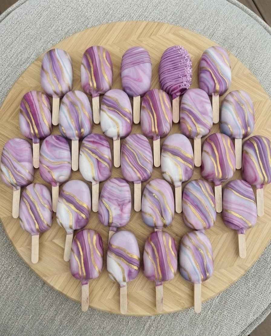 Array of purple, white, and gold marble-patterned cake pops with wooden sticks on a round wooden board.