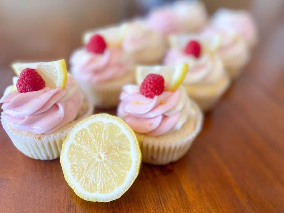 Cupcakes topped with pink frosting, a raspberry, and a lemon wedge, arranged on a wooden surface.